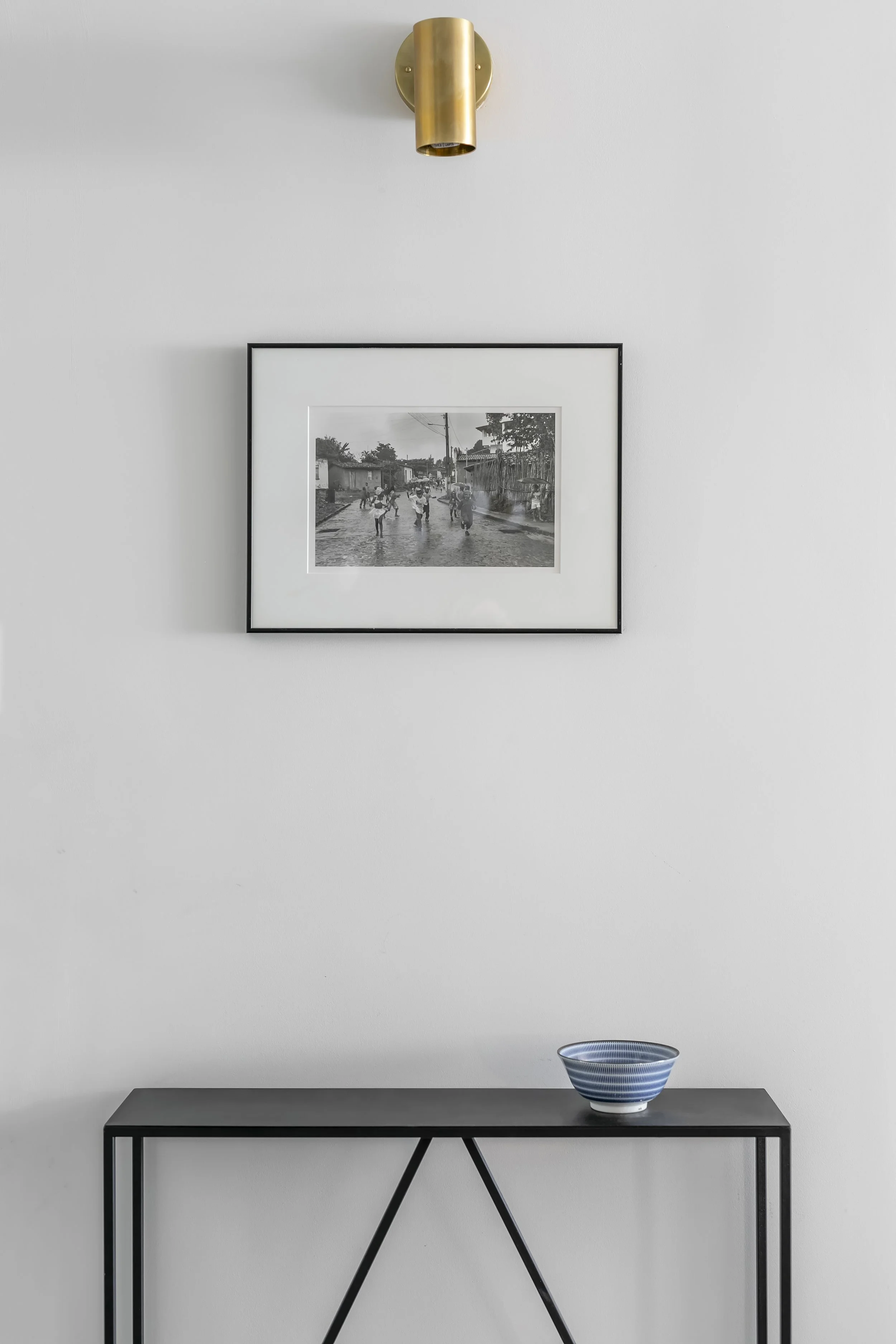 A minimalist interior wall decor with a black and white framed photo, a brass wall sconce above, and a black metal console table with a blue and white striped bowl.