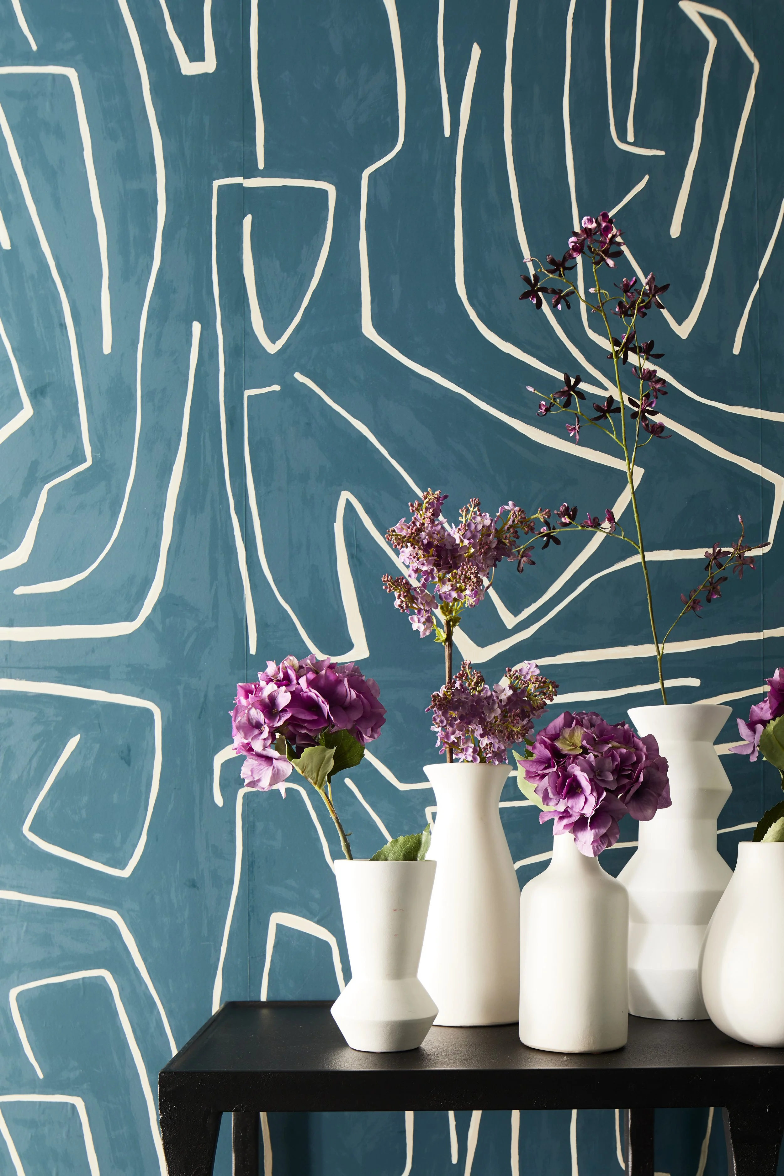 Five white vases with purple and pink flowers on a black table against a blue wall with white abstract line art.