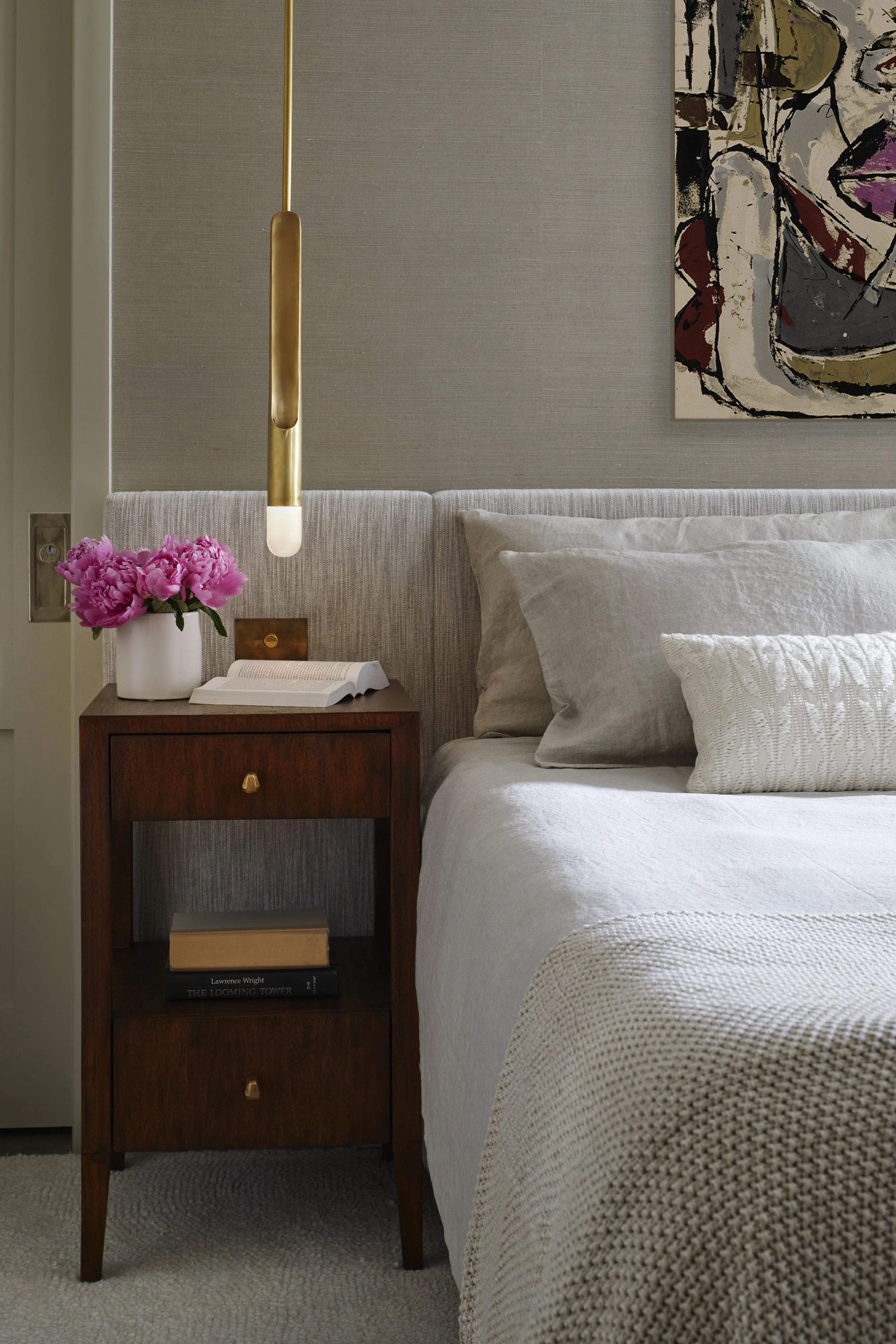 A bedroom with a beige headboard, a wooden nightstand with books and a white book open, a pink flower arrangement, a white textured pillow, a grey bedspread, a hanging gold light fixture, and a colorful abstract painting on the wall.
