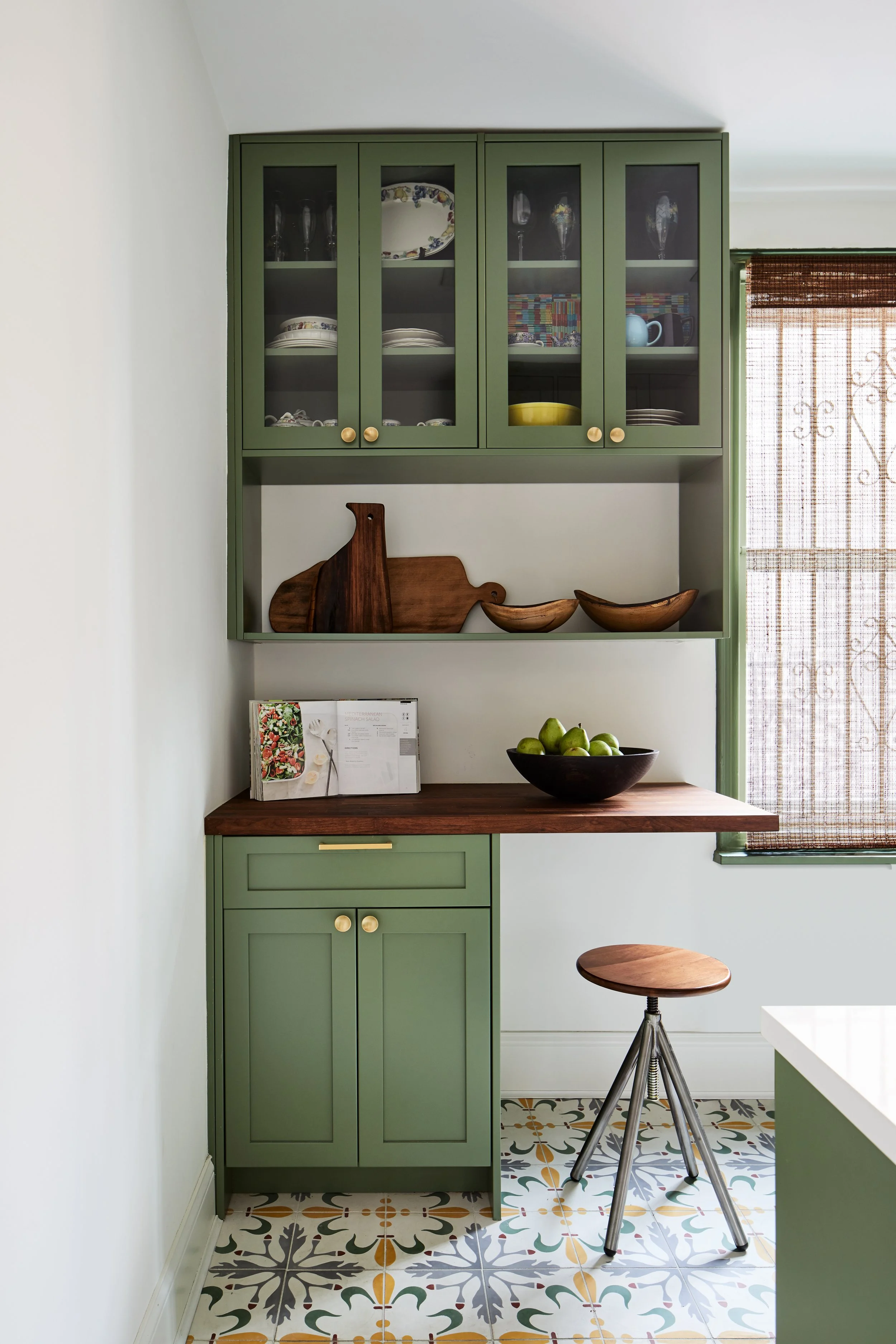 A kitchen corner with green cabinetry, a wooden countertop, a stool, and a window with a woven shade. The upper cabinets display glassware and dishes, while the lower includes a bowl of green apples on the counter.