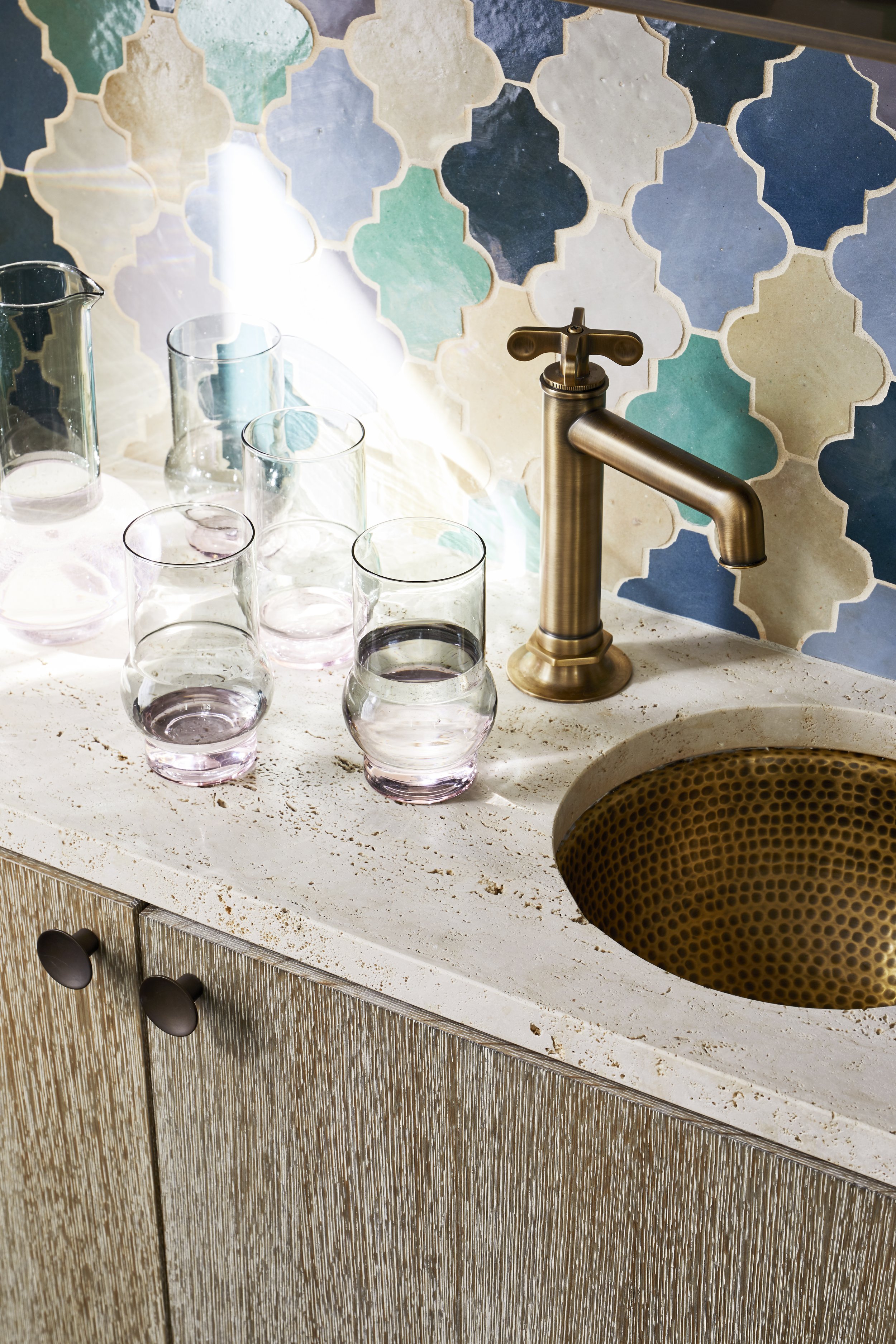A white kitchen countertop with a gold-colored sink, a gold faucet, and several glasses of water. The backsplash features a pattern of blue, green, beige, and black tiles.