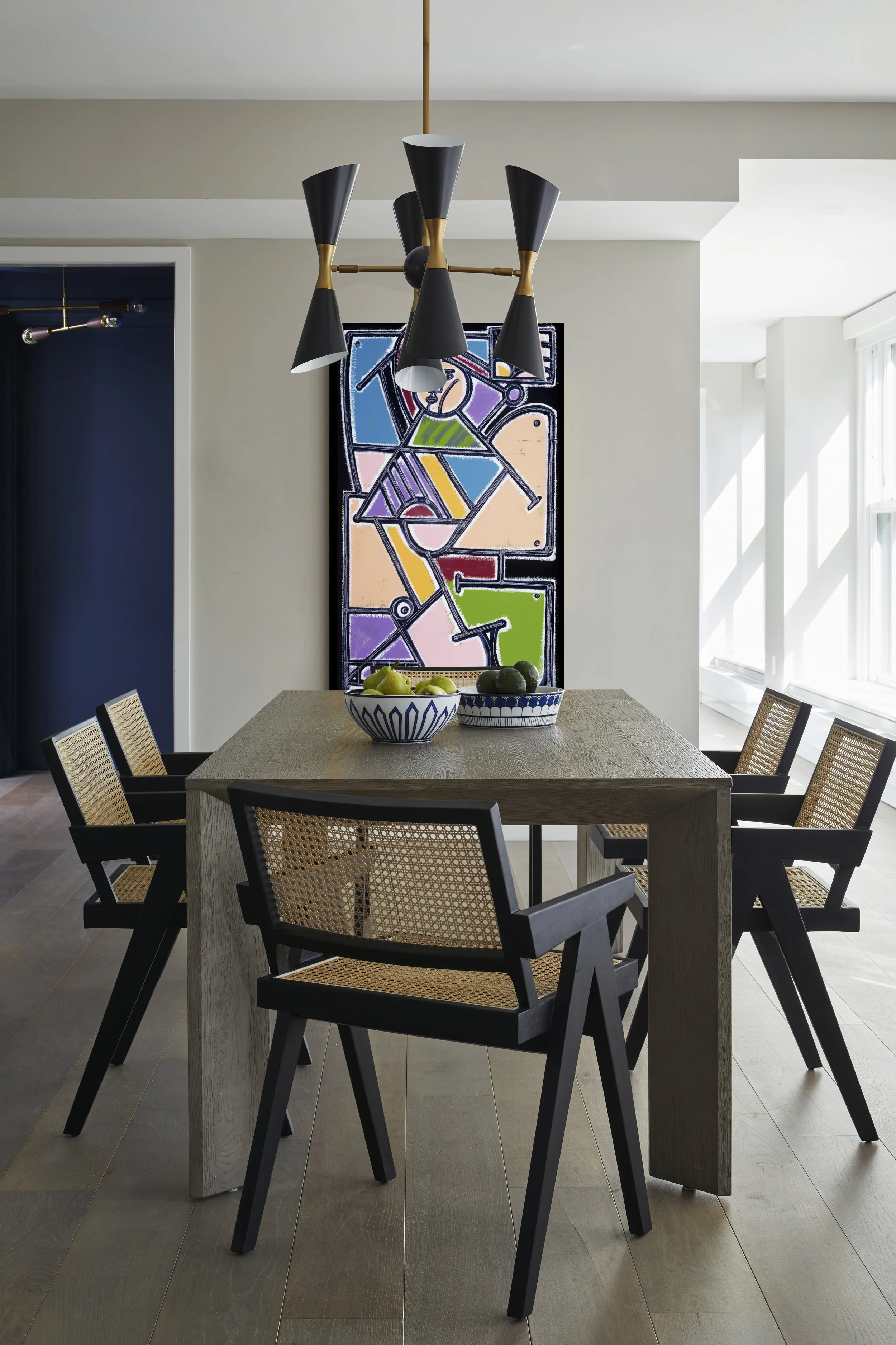Modern dining room with black and wood chairs around a rectangular wooden table. A black and gold chandelier hangs above. Two bowls with fruit sit on the table, and a colorful abstract art piece is on the wall.