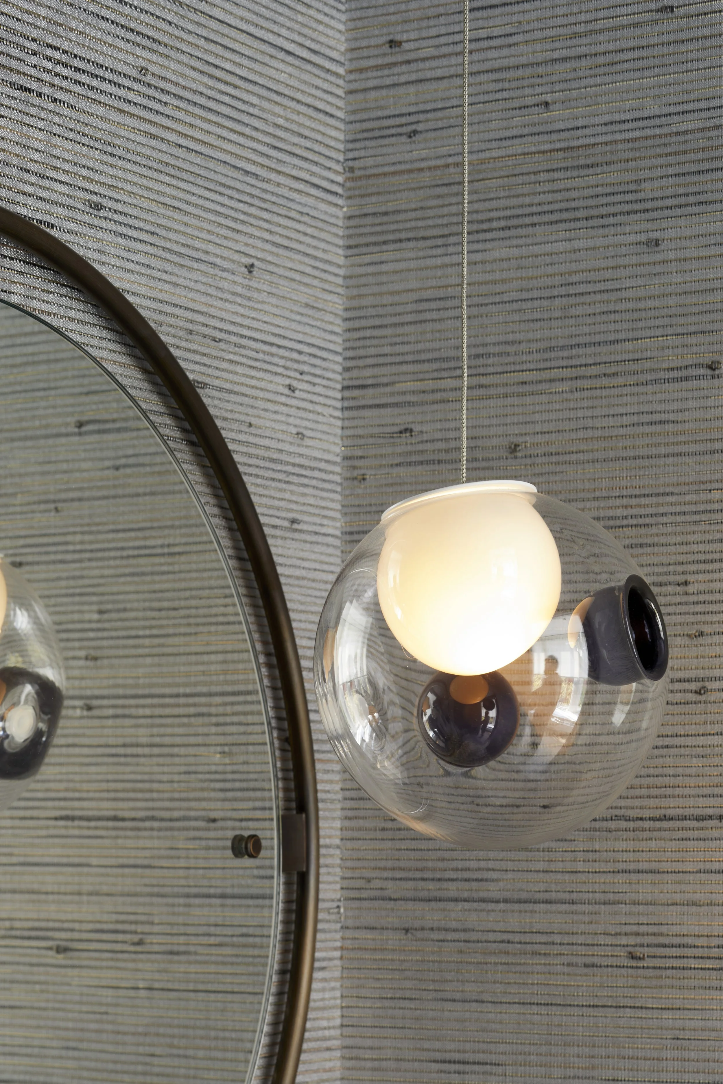 A modern wall-mounted light fixture with a glass globe shade illuminating a textured wall with horizontal lines, reflected in a round mirror.