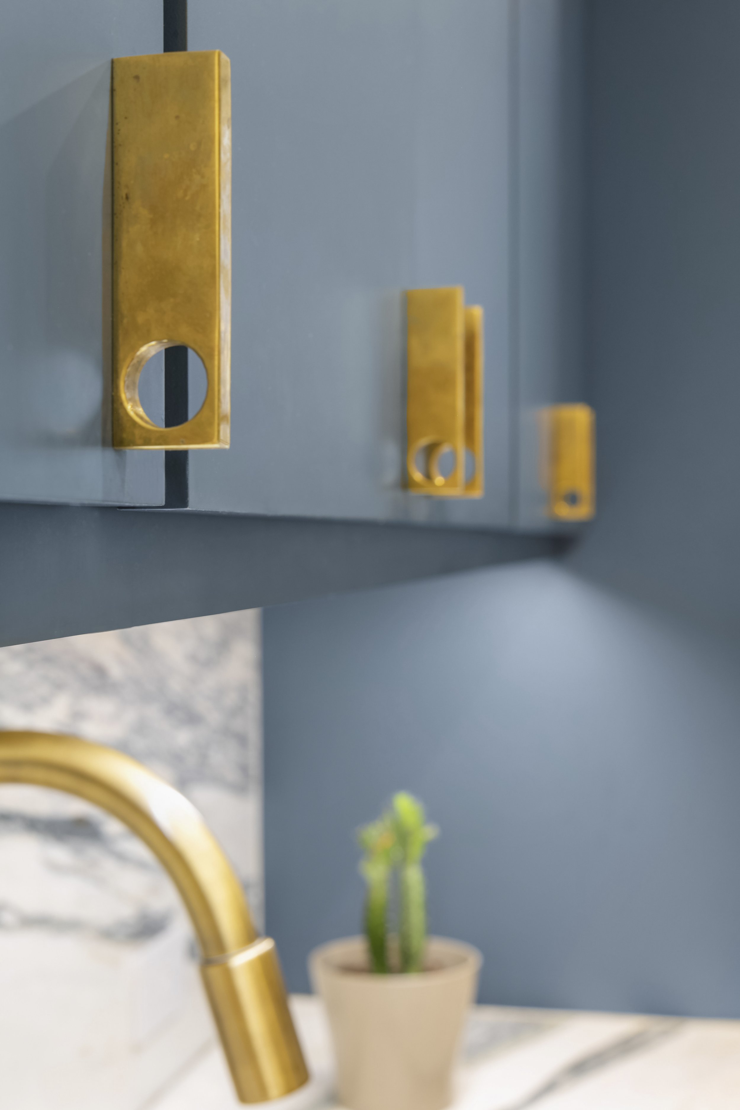 Close-up of blue kitchen cabinets with gold handles, with a blurred potted cactus and gold kitchen faucet in the background.