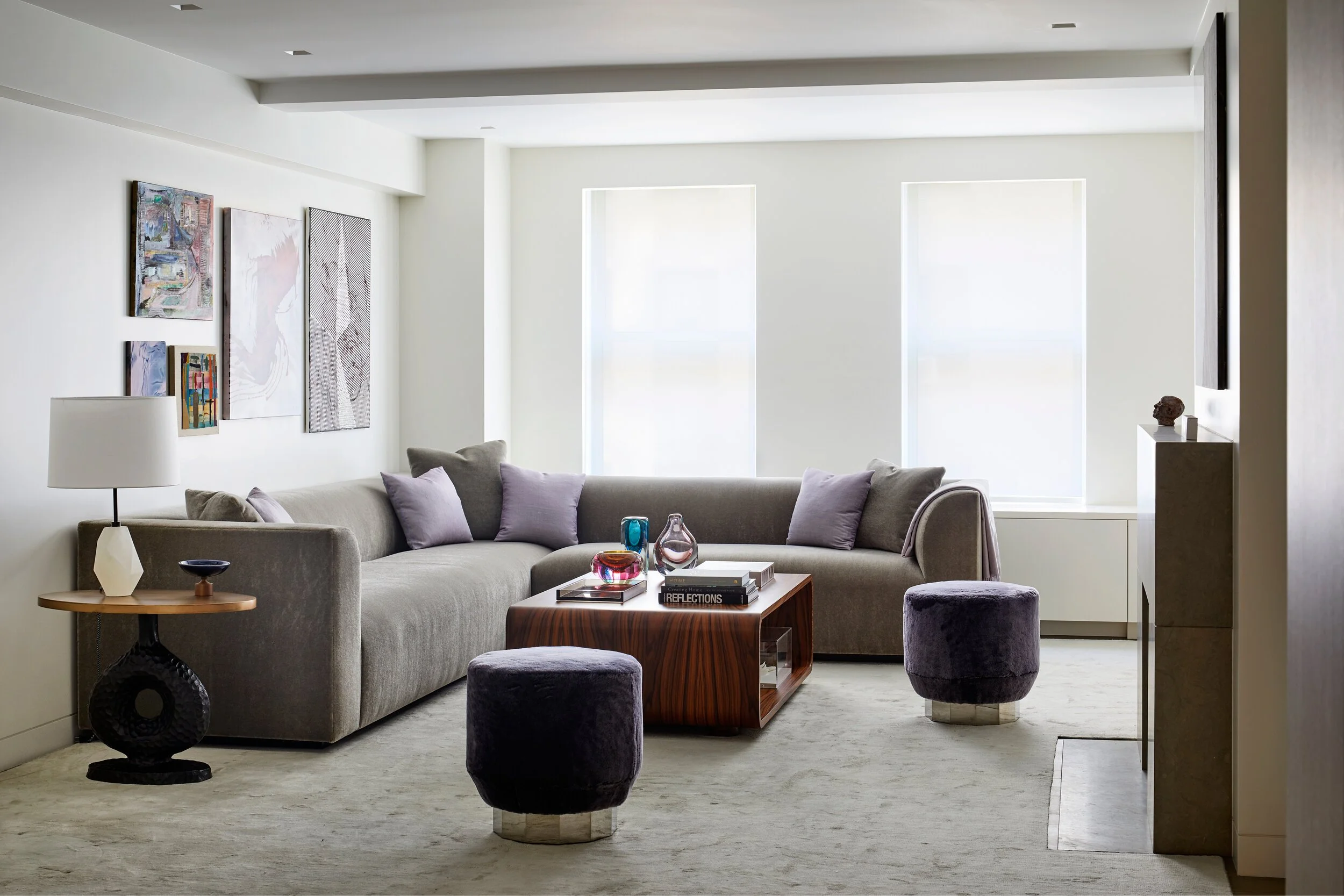 Modern living room with a beige sectional sofa, purple pillows, two purple ottomans, a wooden coffee table with books and glass vases, a side table with a lamp, abstract art on the wall, and large windows with sheer shades.
