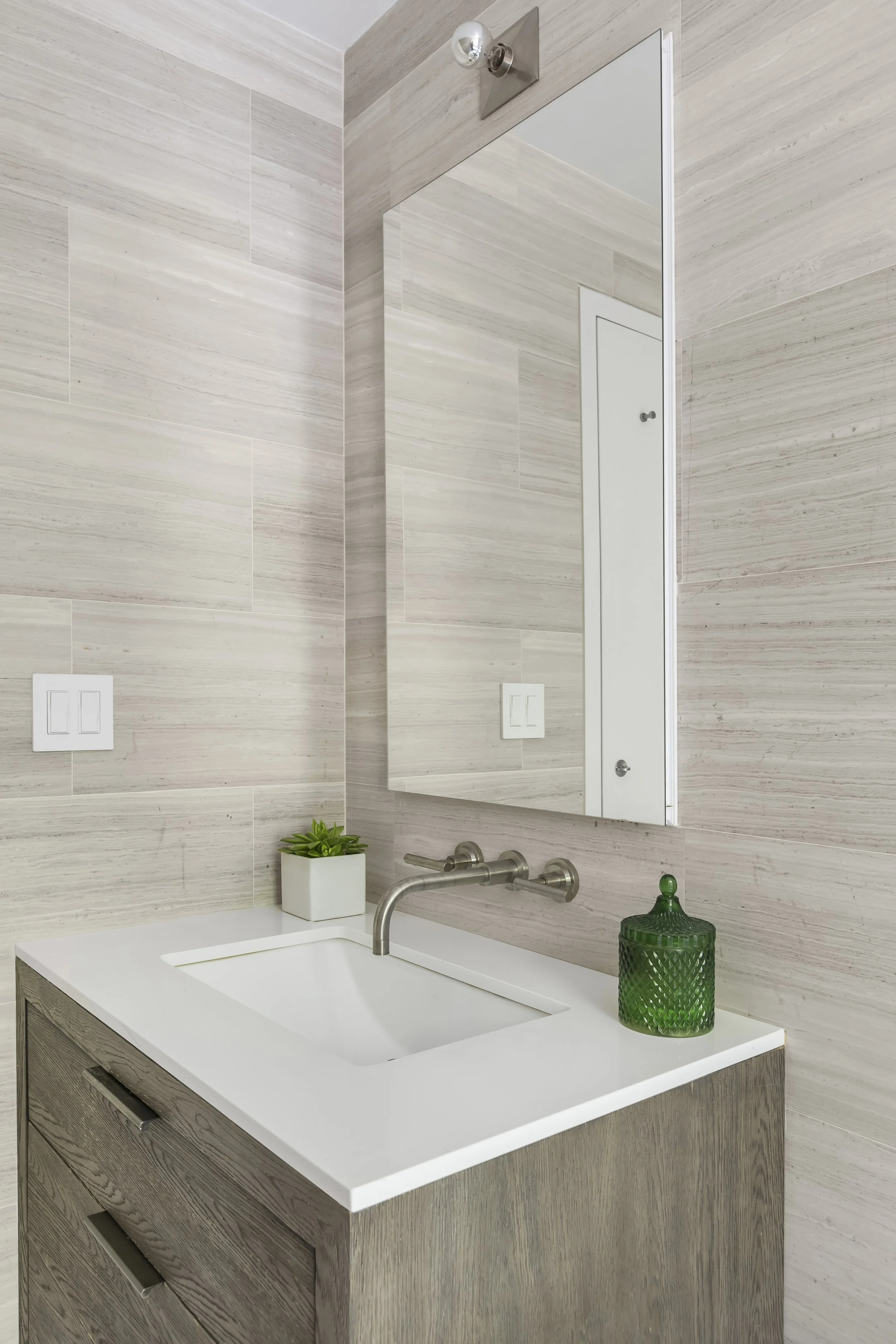 A modern bathroom sink with a light wood vanity, a white countertop, a rectangular mirror, a small potted plant, a green glass jar with a lid, and a wall-mounted faucet.