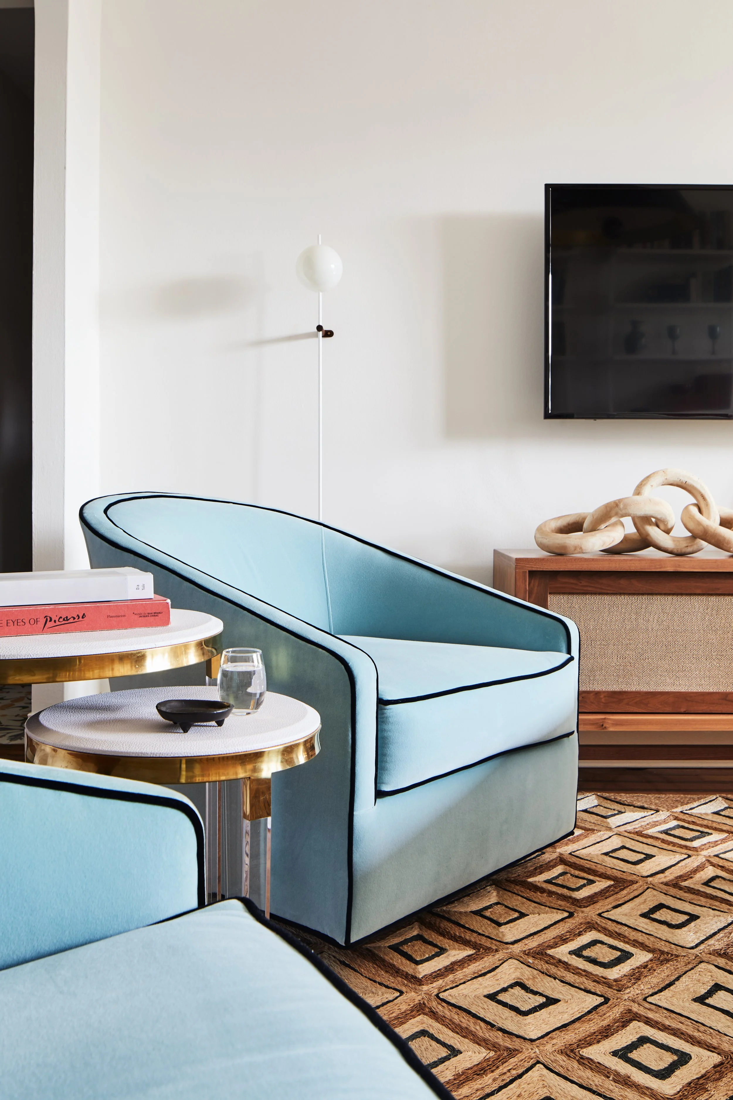 Living room with light blue armchair, side tables, a glass of water, a bowl, a wall-mounted TV, a wooden console with sculpture, wall lamp, and patterned rug.