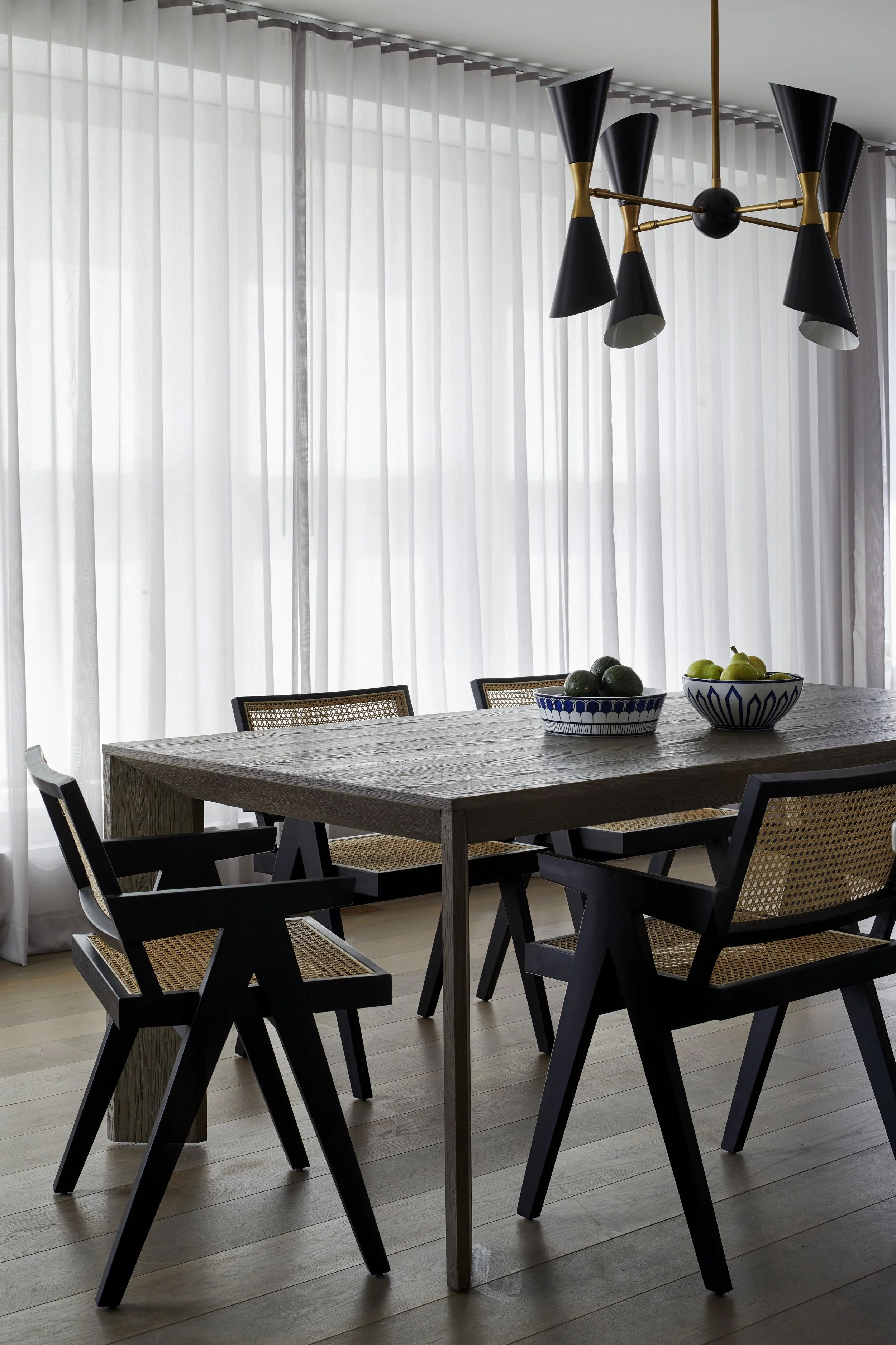 A dining room with a wooden table, black chairs with woven seats, and a modern chandelier hanging above. There are bowls with green and dark fruits on the table, and sheer white curtains covering large windows.