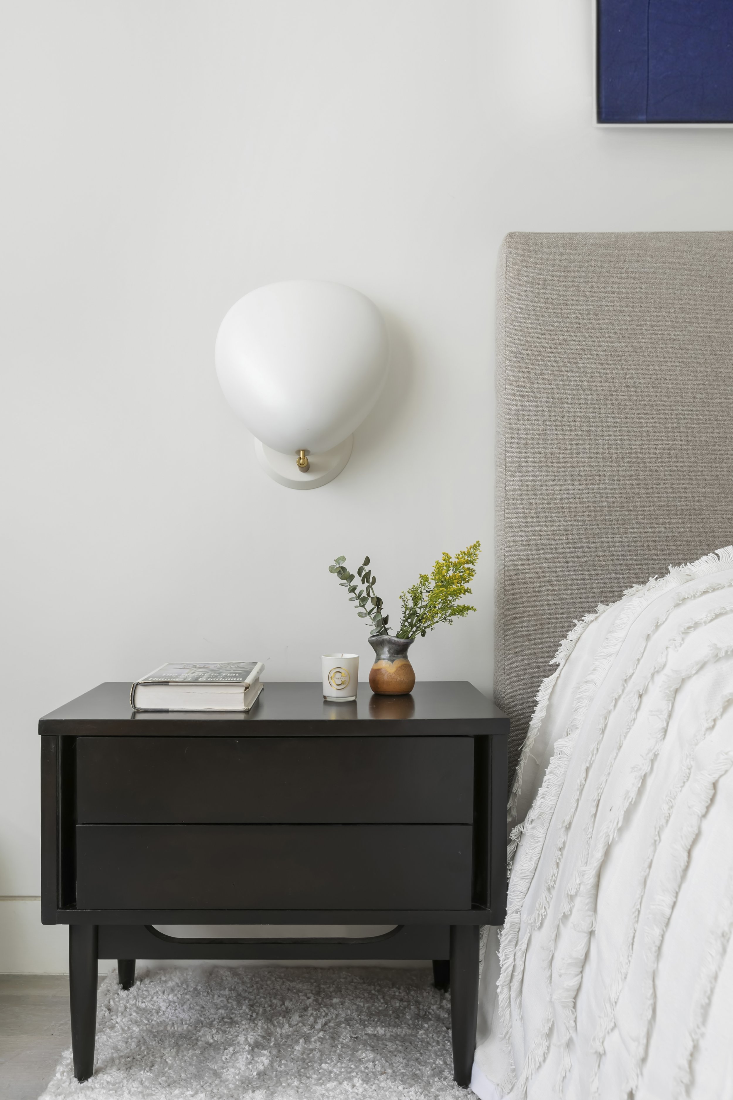 A bedroom with a white wall, a wall-mounted white sconce, a black nightstand, a book, a small vase with greenery, and a white bed with textured bedding.