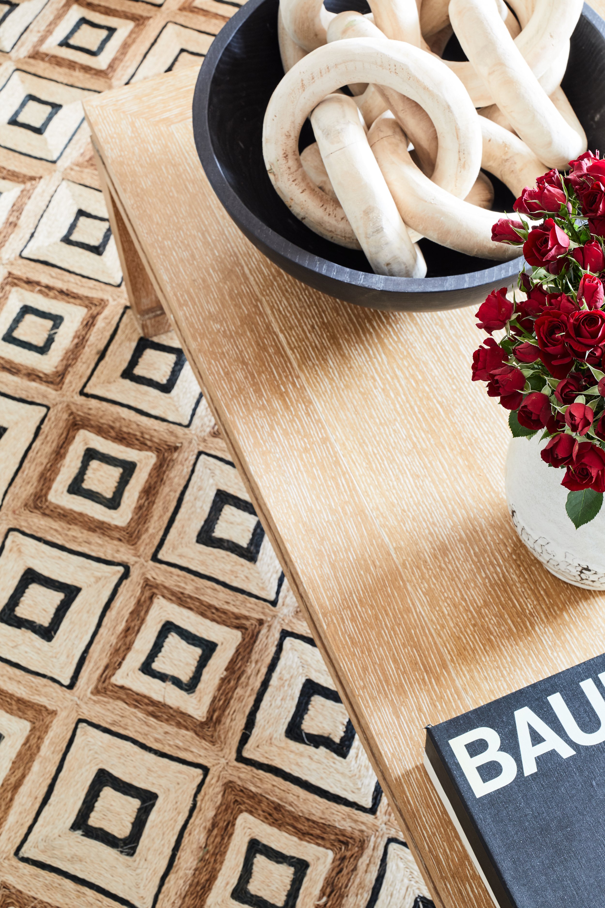 A wooden table with a black bowl filled with white, thick ropes, a vase of red roses, and a black and white book. The table is on a geometric patterned rug with square motifs in shades of beige, black, and brown.
