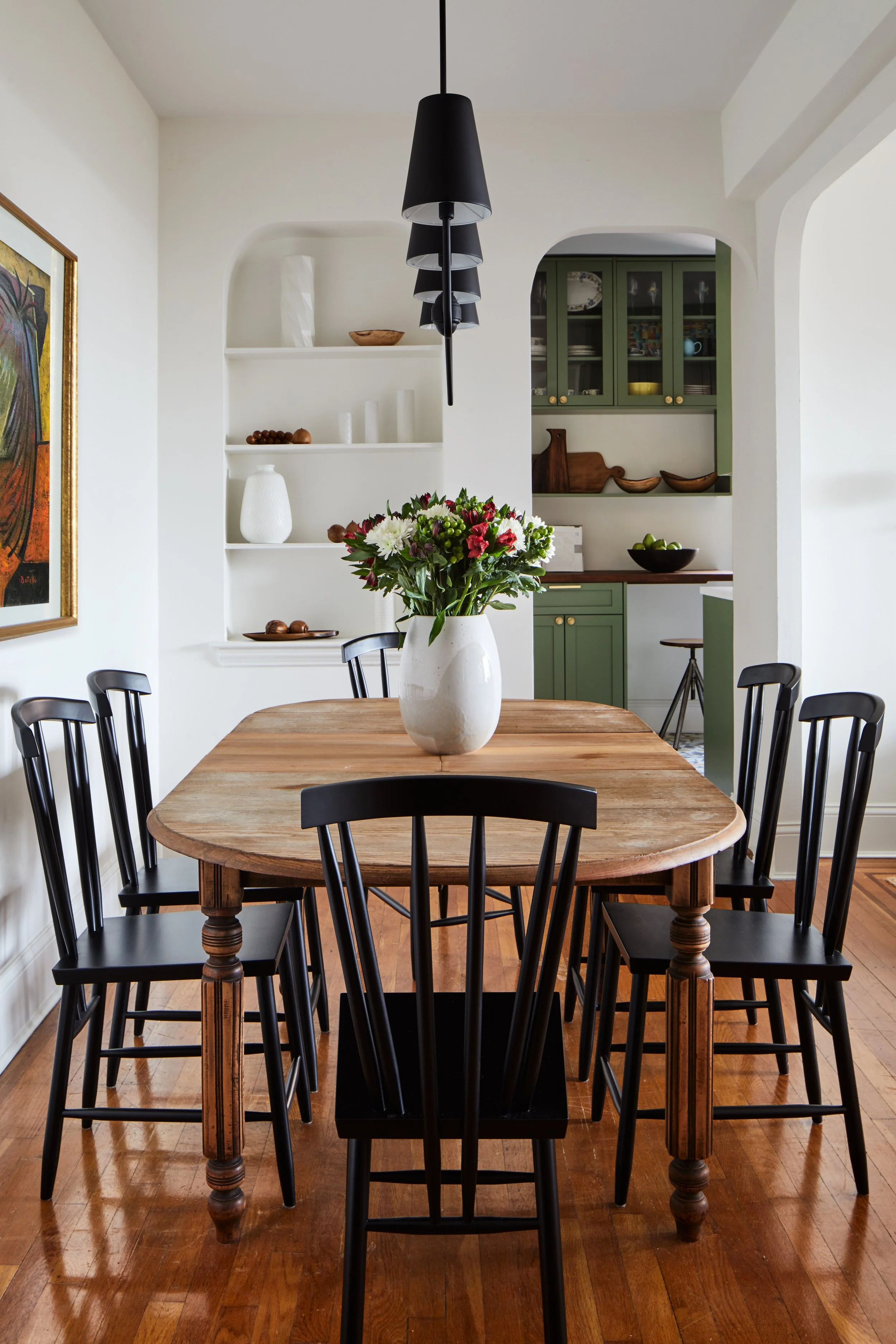 A dining room with a wooden table, black chairs, a white vase with flowers, a hanging black pendant light, and a kitchen with green cabinets in the background.