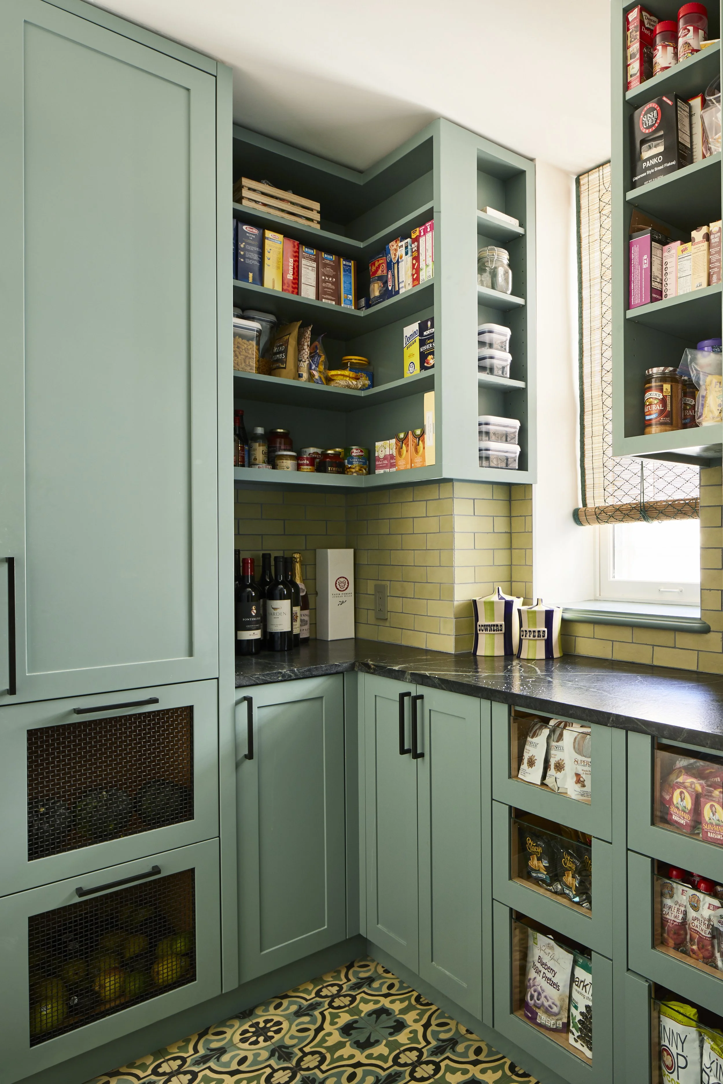 A cozy kitchen with light green cabinets, black countertops, and a patterned tile floor. There are open shelves with food and spice containers, a window with a bamboo shade, and yellow brick backsplash.