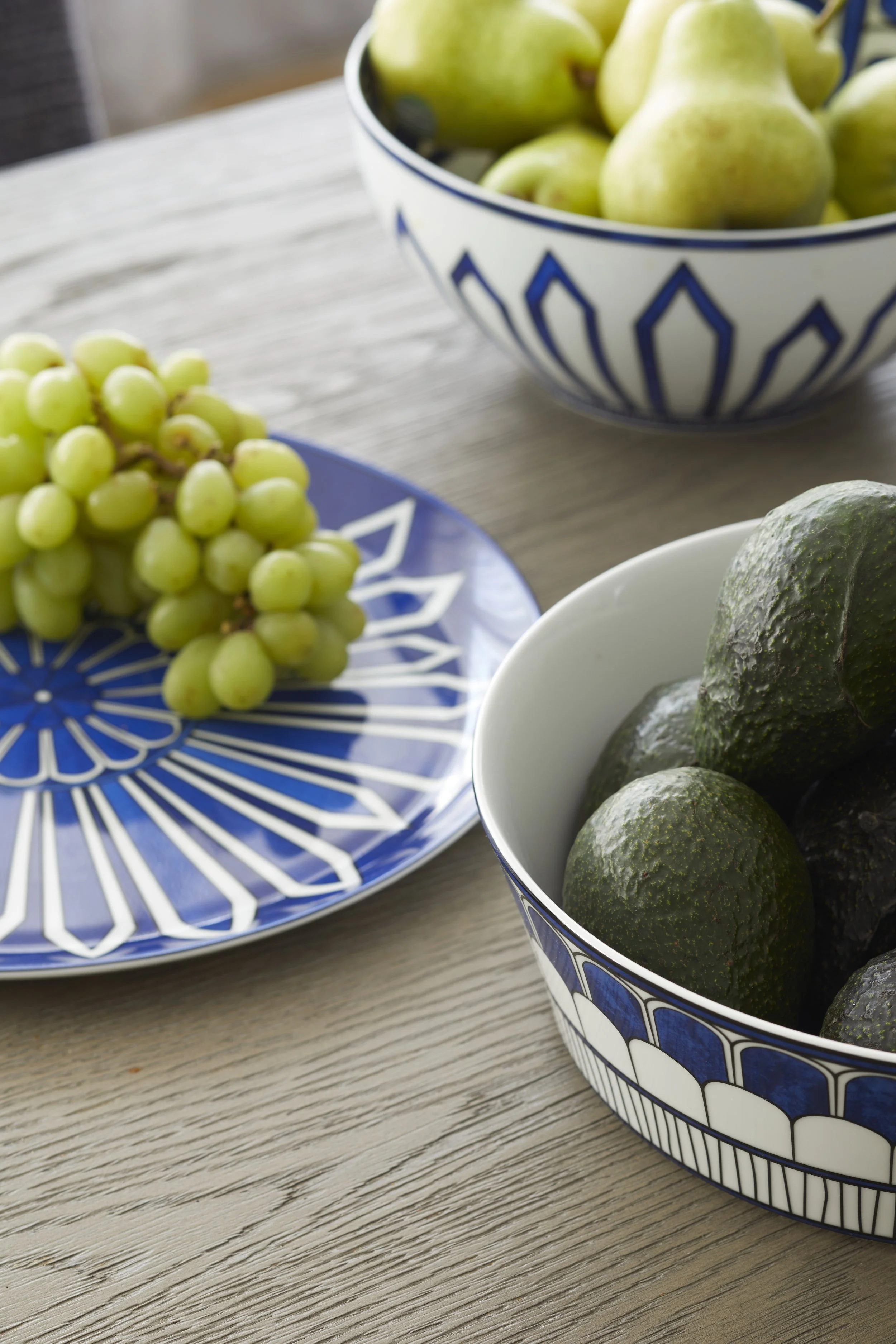 A table with a bunch of green grapes on a blue and white patterned plate, a bunch of green pears in a white bowl with blue geometric designs, and several avocados in another white bowl with blue designs.