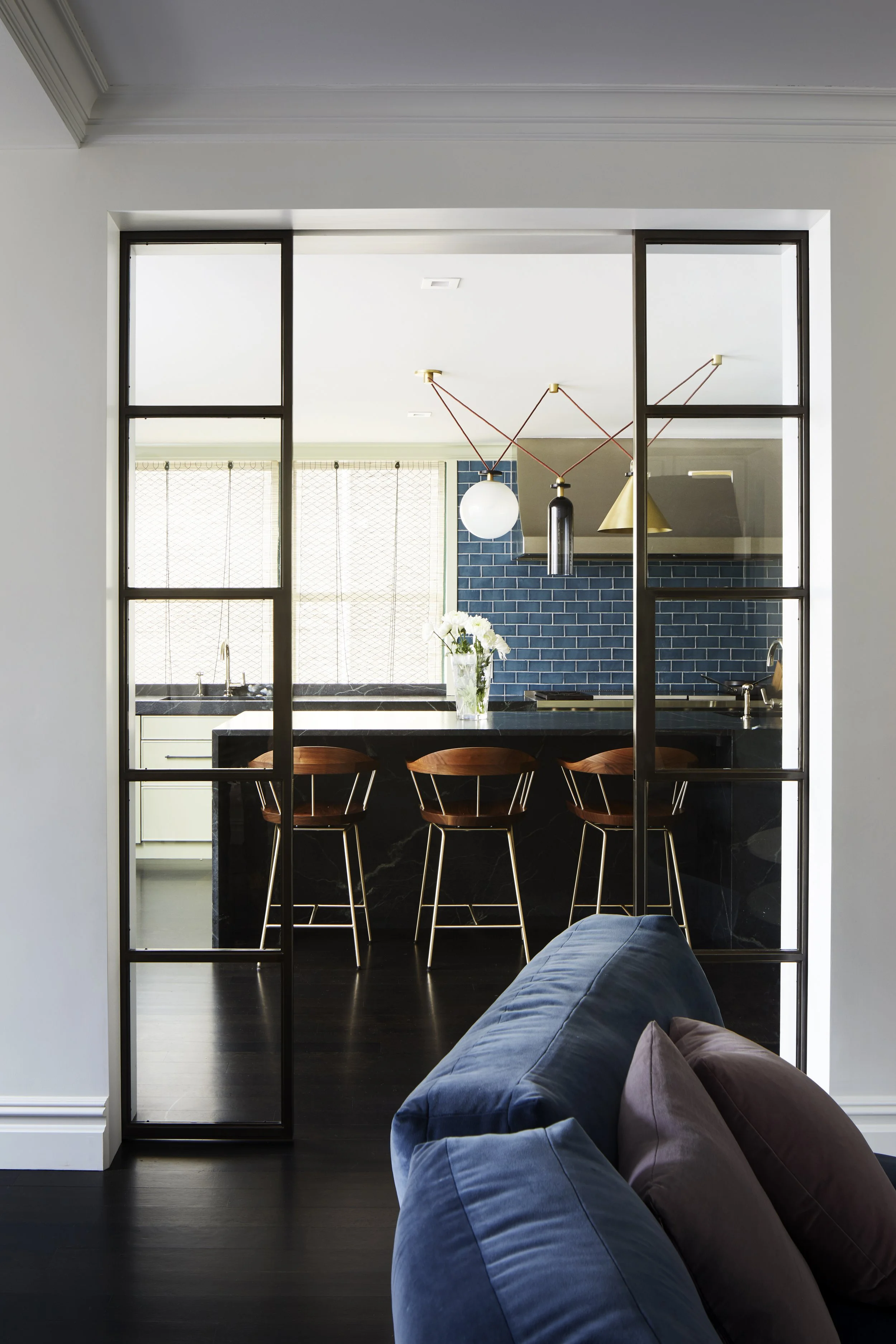 View through glass panel of modern kitchen with blue tile backsplash, dark countertops, three wooden bar stools, and pendant lights, from living room with blue and brown curtains.