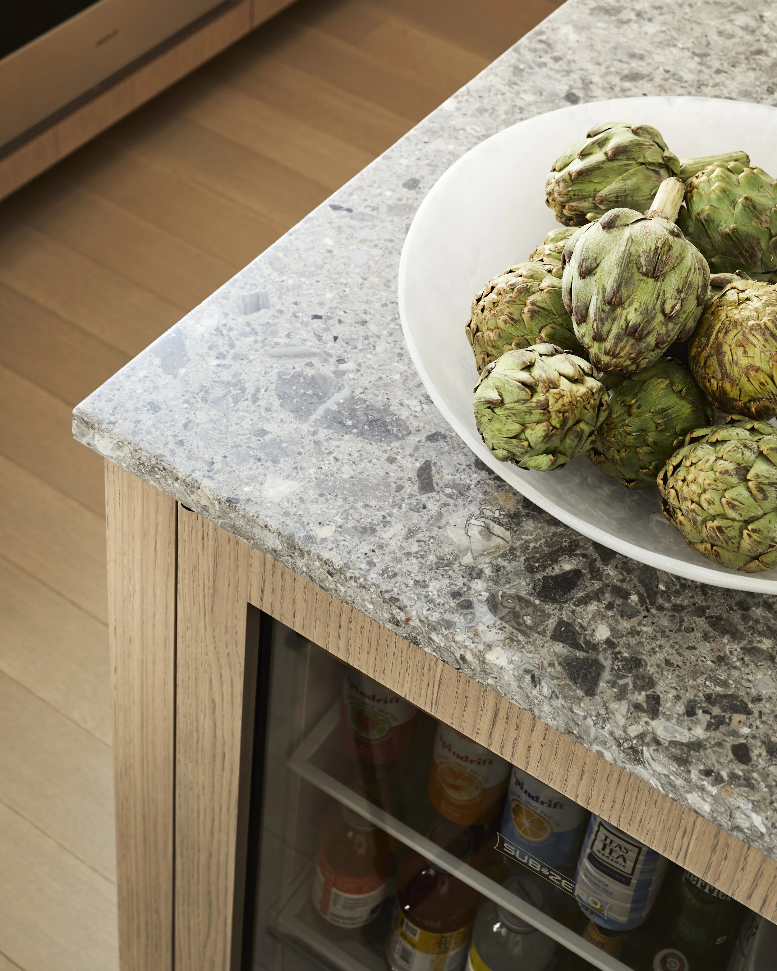 Bunch of fresh artichokes on a white plate on a kitchen countertop with open cabinet below containing bottles.