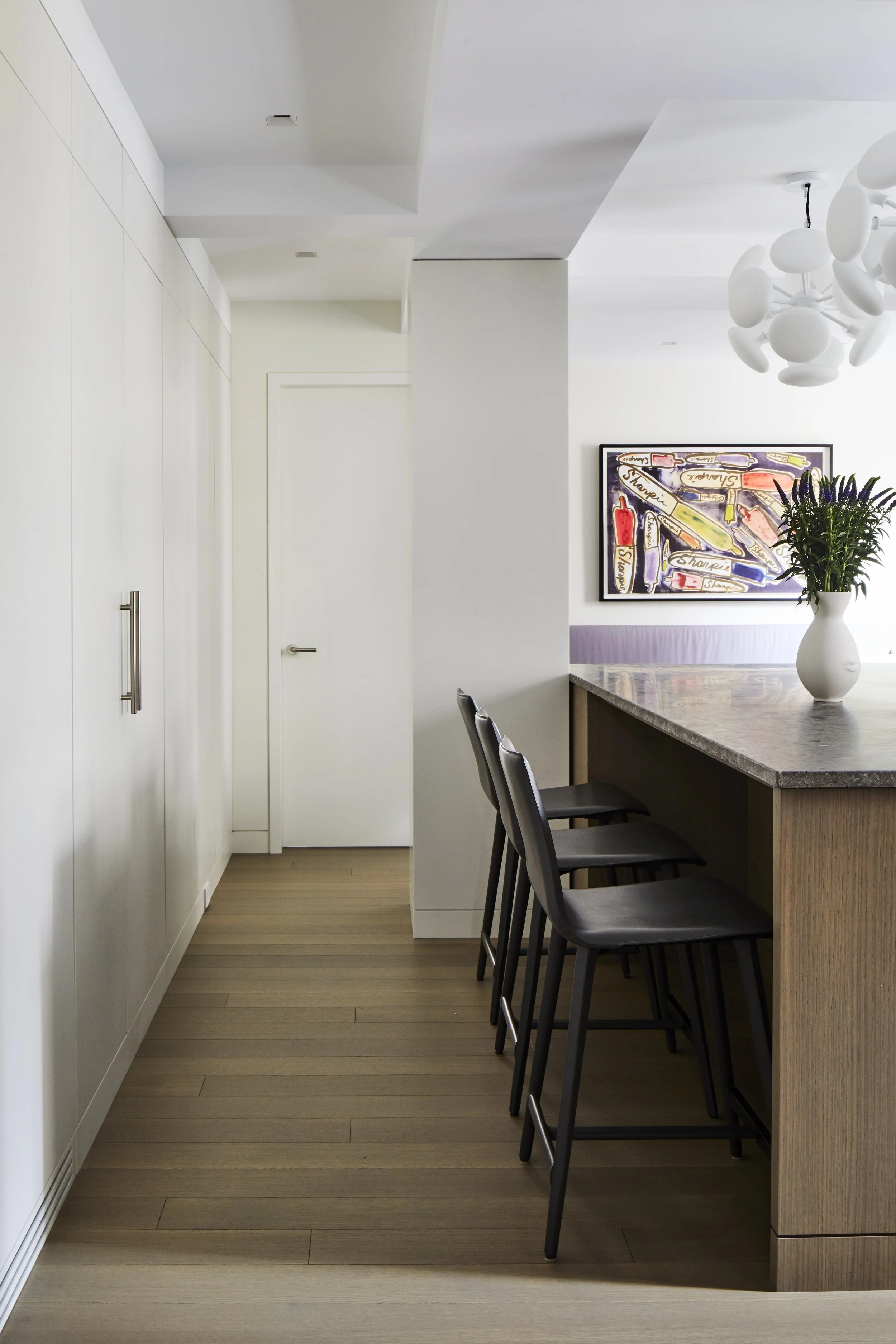 Modern kitchen with white cabinets, a wooden island with bar stools, a vase of purple flowers, and colorful artwork on the wall.