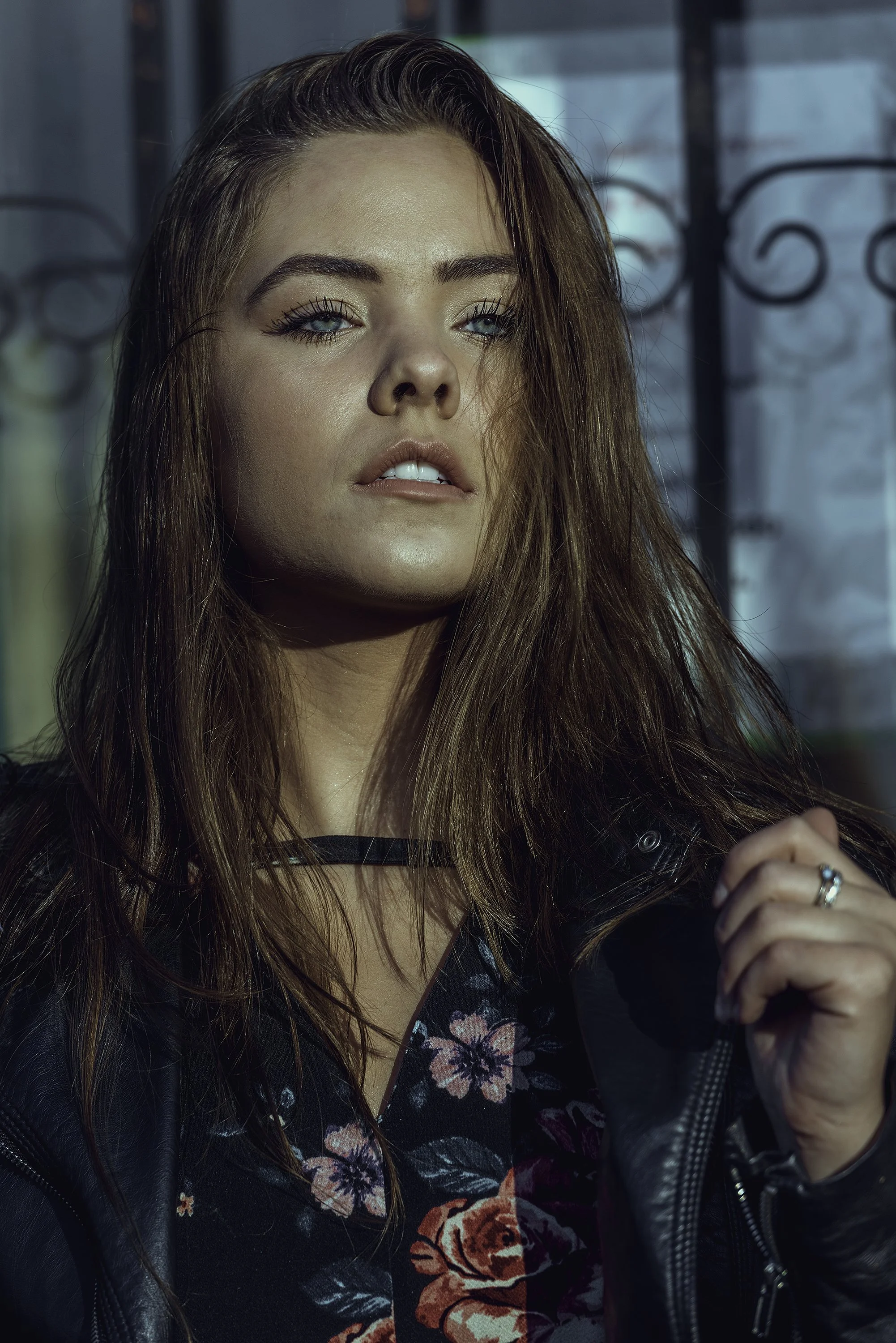 Outdoor fashion portrait of a woman with long brown hair wearing a dark floral outfit and black leather jacket, photographed against an architectural background.