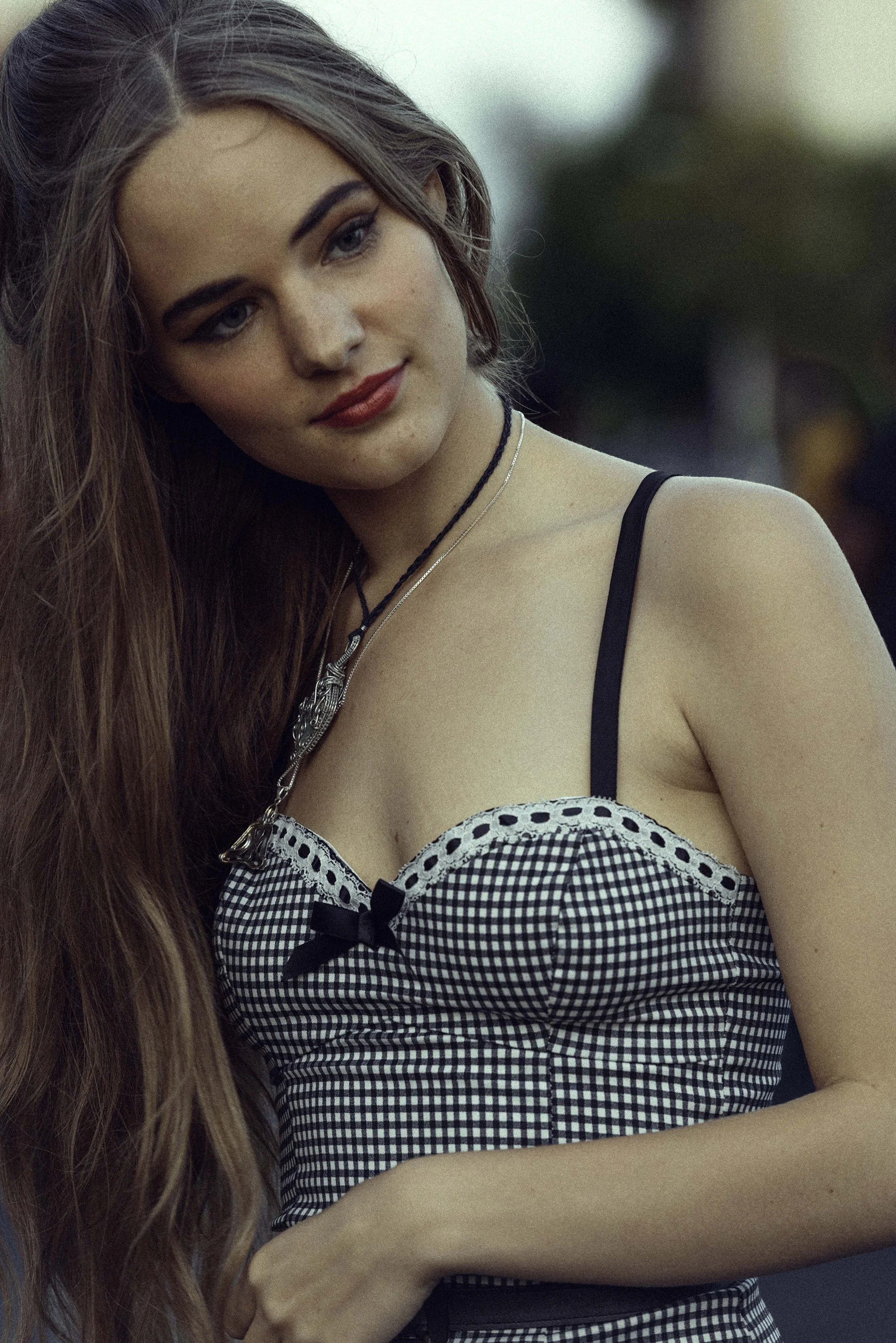 Outdoor fashion portrait of a woman with long brown hair wearing a black-and-white checkered top with lace detailing and layered necklaces, against soft greenery.
