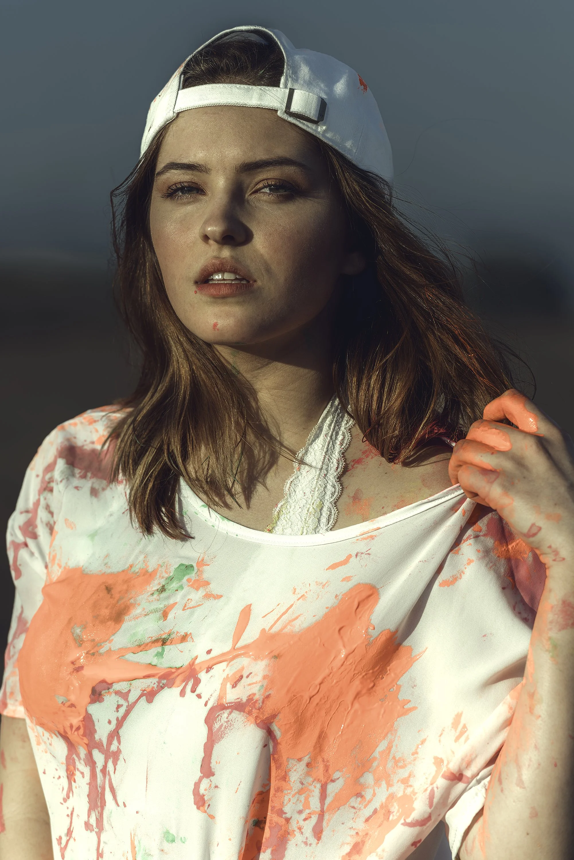 Outdoor fashion portrait of a woman wearing a backward white baseball cap and a white shirt with colorful paint splashes, against a softly blurred background.