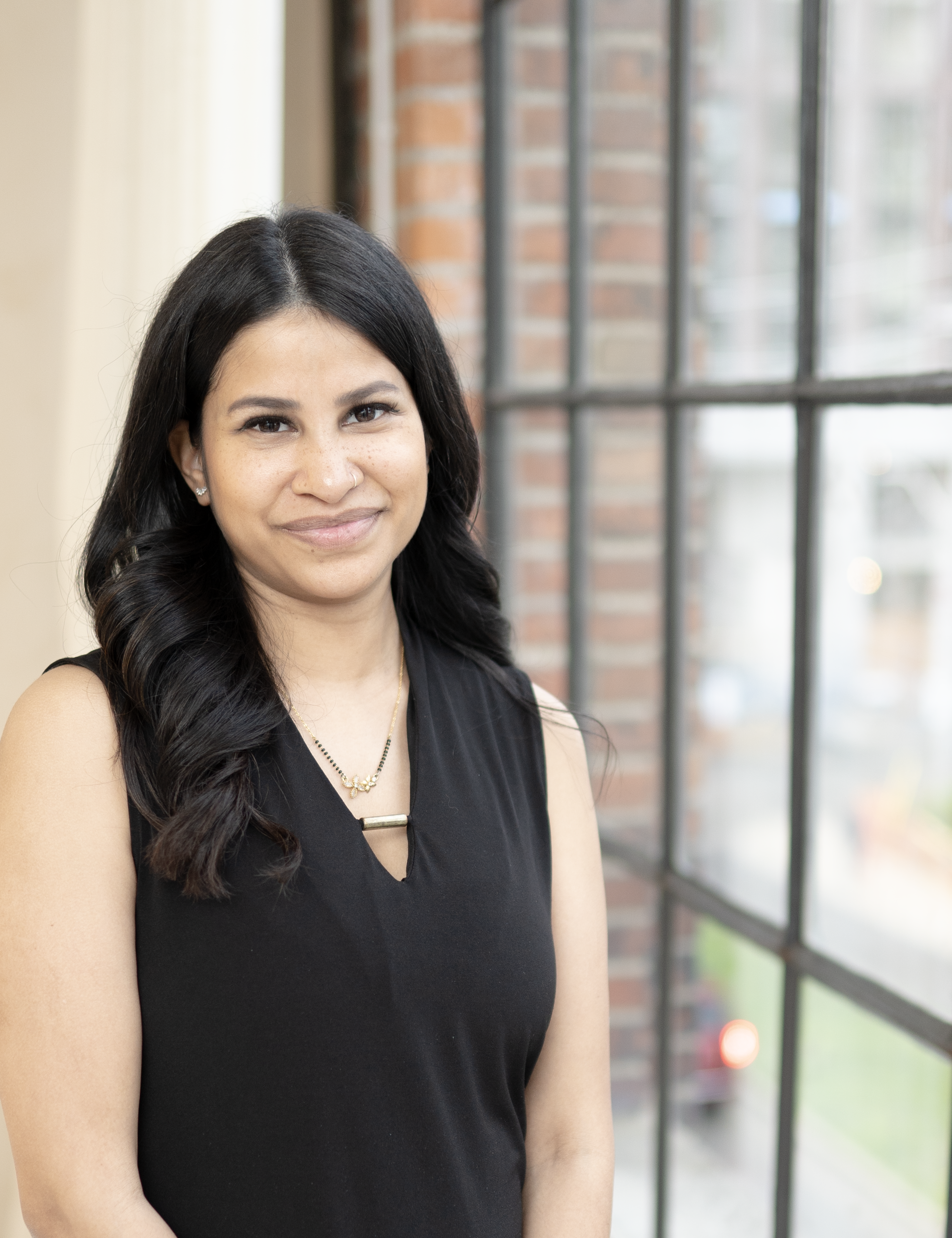 A woman with long black hair, wearing a black sleeveless top, standing in front of a large window with a brick building outside.