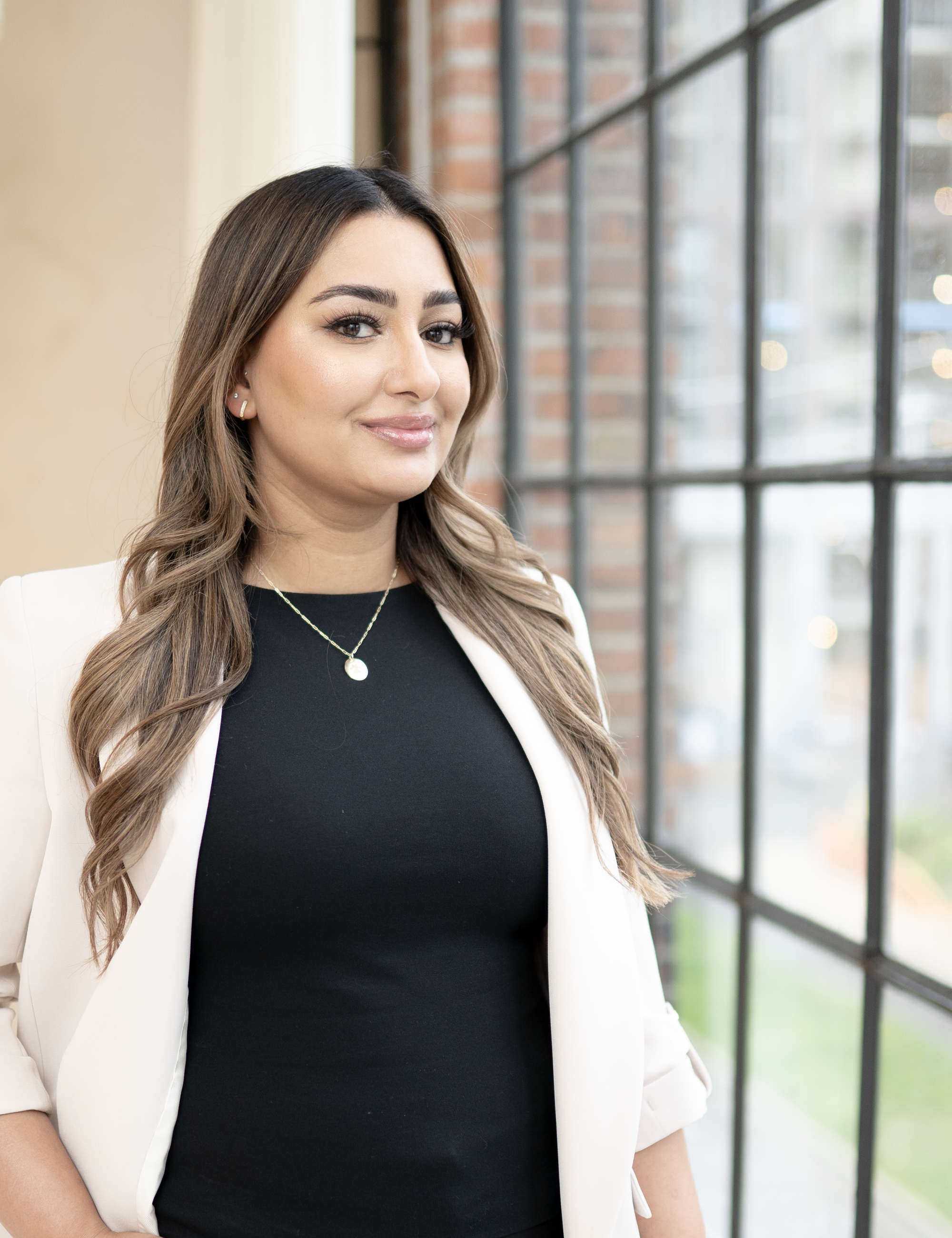 A woman with long brown hair, wearing a white blazer over a black top, standing near a large window with brick and glass panes.
