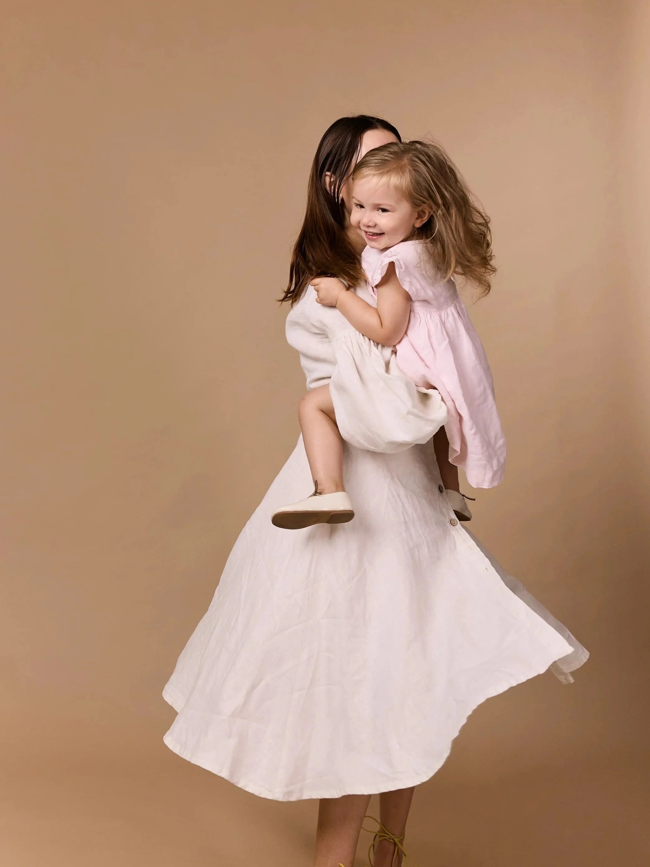 A self portrait studio young woman with long red hair, blue eyes, and freckles, sitting sideways on a black stool against an orange background, wearing a white sleeveless top and white lace skirt, with a serious expression and wearing drop earrings.