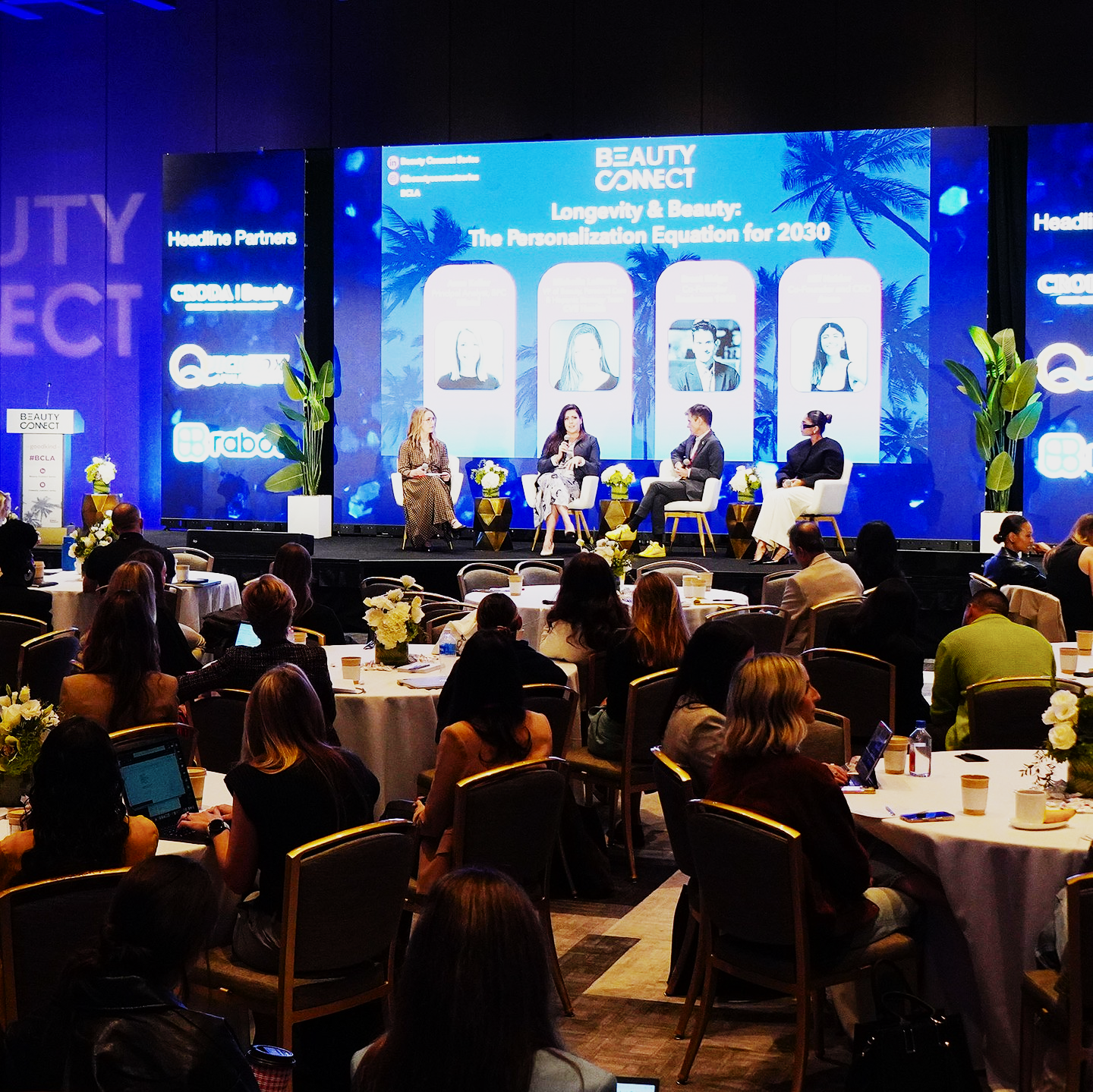 A panel discussion at a conference with four women seated on stage, microphones in hand, and an audience of diverse attendees watching. The background displays a blue screen with event branding and images of four women.