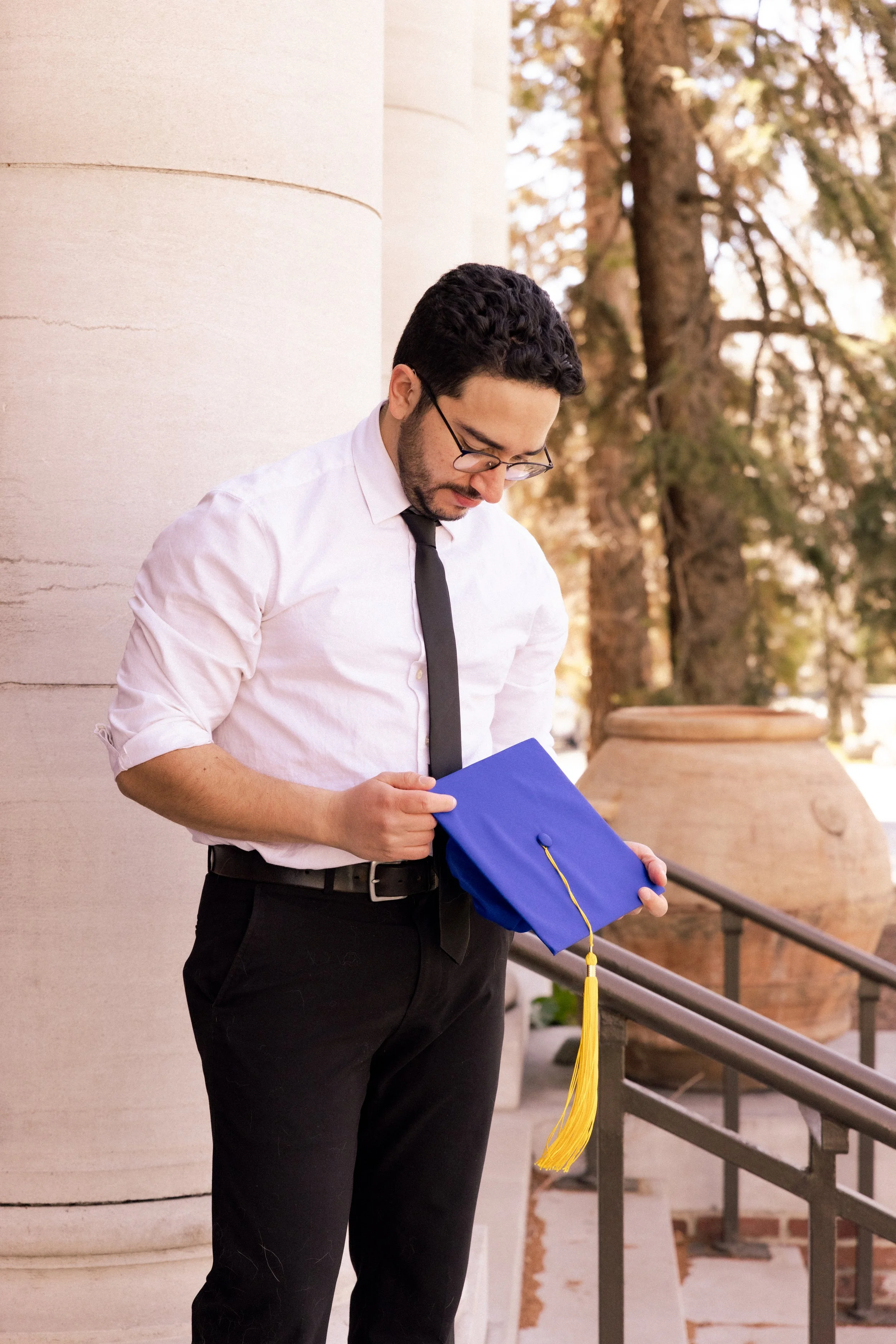 A man in a white shirt, black tie, and glasses stands outdoors, holding a blue graduation cap with a yellow tassel, looking down at it.