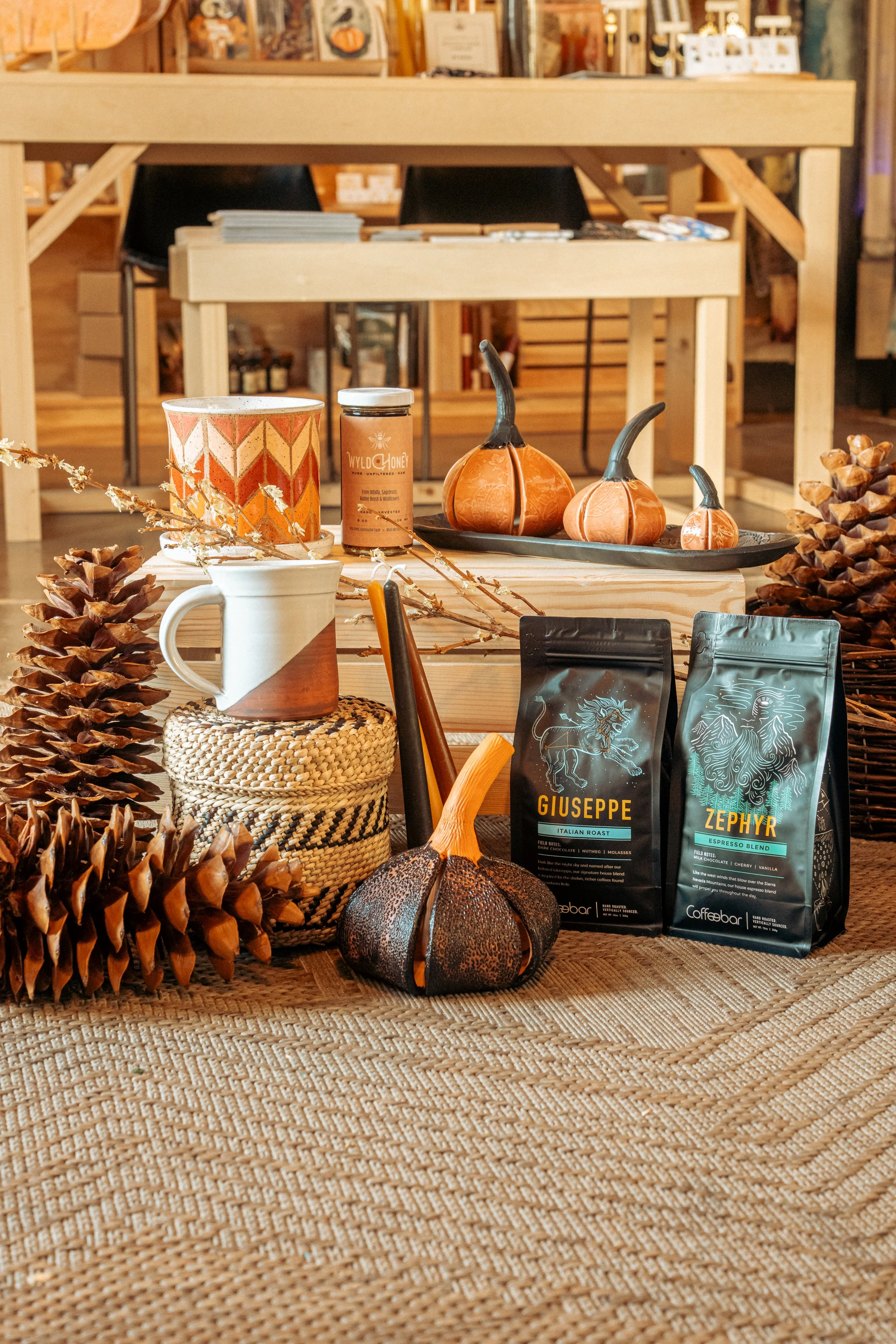 Autumn-themed coffee display with pumpkins, pinecones, dried branches, and coffee bags on a woven tablecloth.