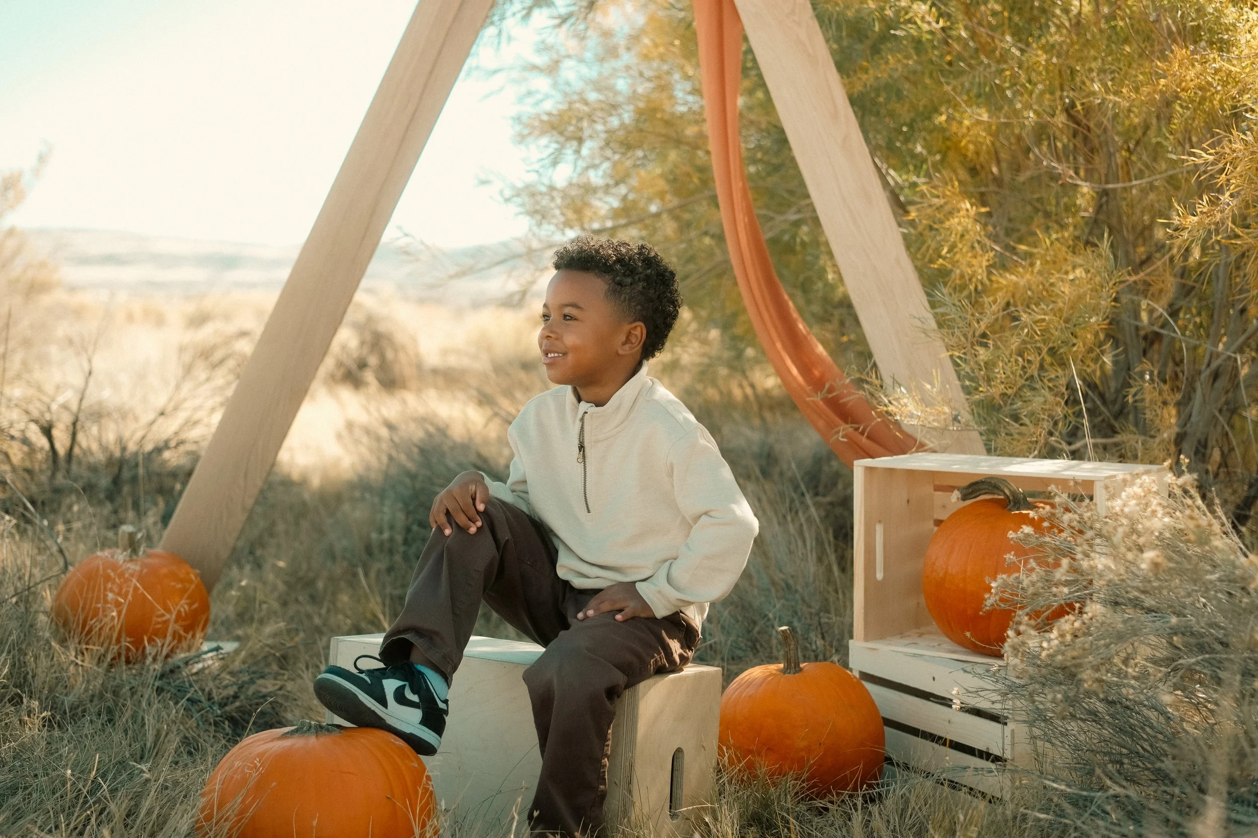 Smiling boy sitting on a wooden box outdoors surrounded by pumpkins and fall foliage