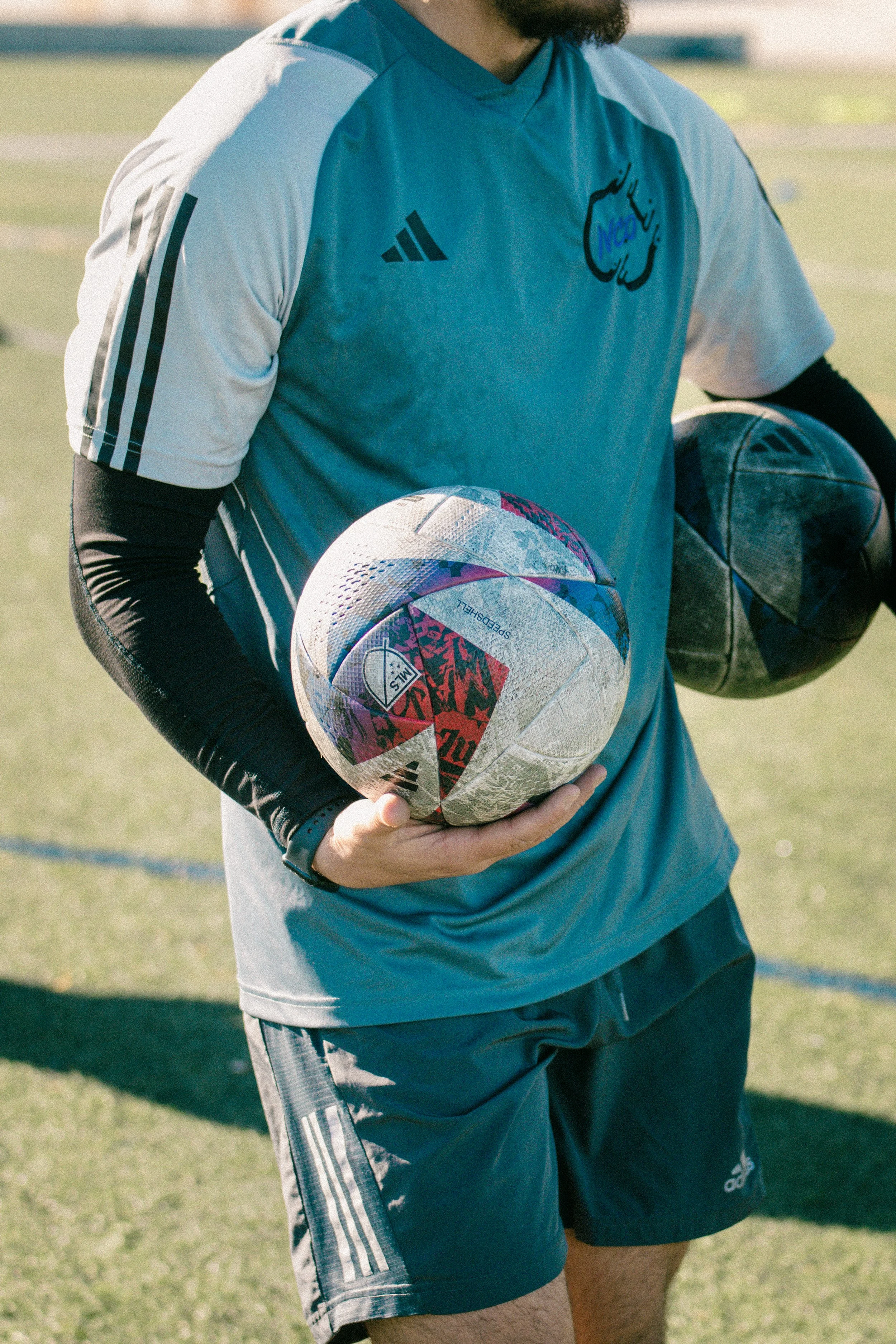 A person holding two soccer balls, standing on a field wearing athletic shorts and a blue and white jersey.