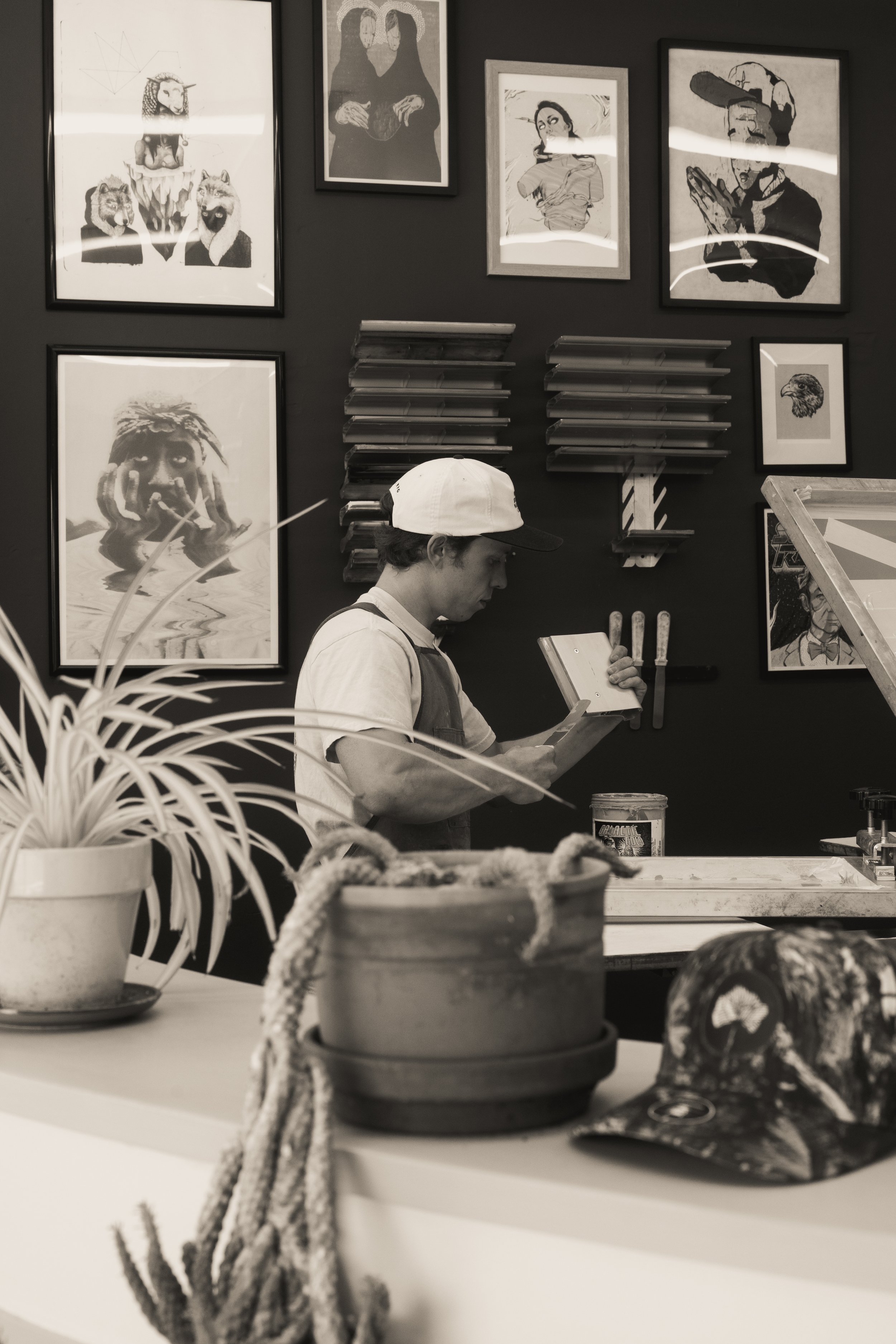 A young man is standing behind a counter in an art gallery or studio, looking at a tablet or notebook. The wall behind him features multiple framed artworks, including portraits and illustrations. There are potted plants and a cap on the counter in the foreground.