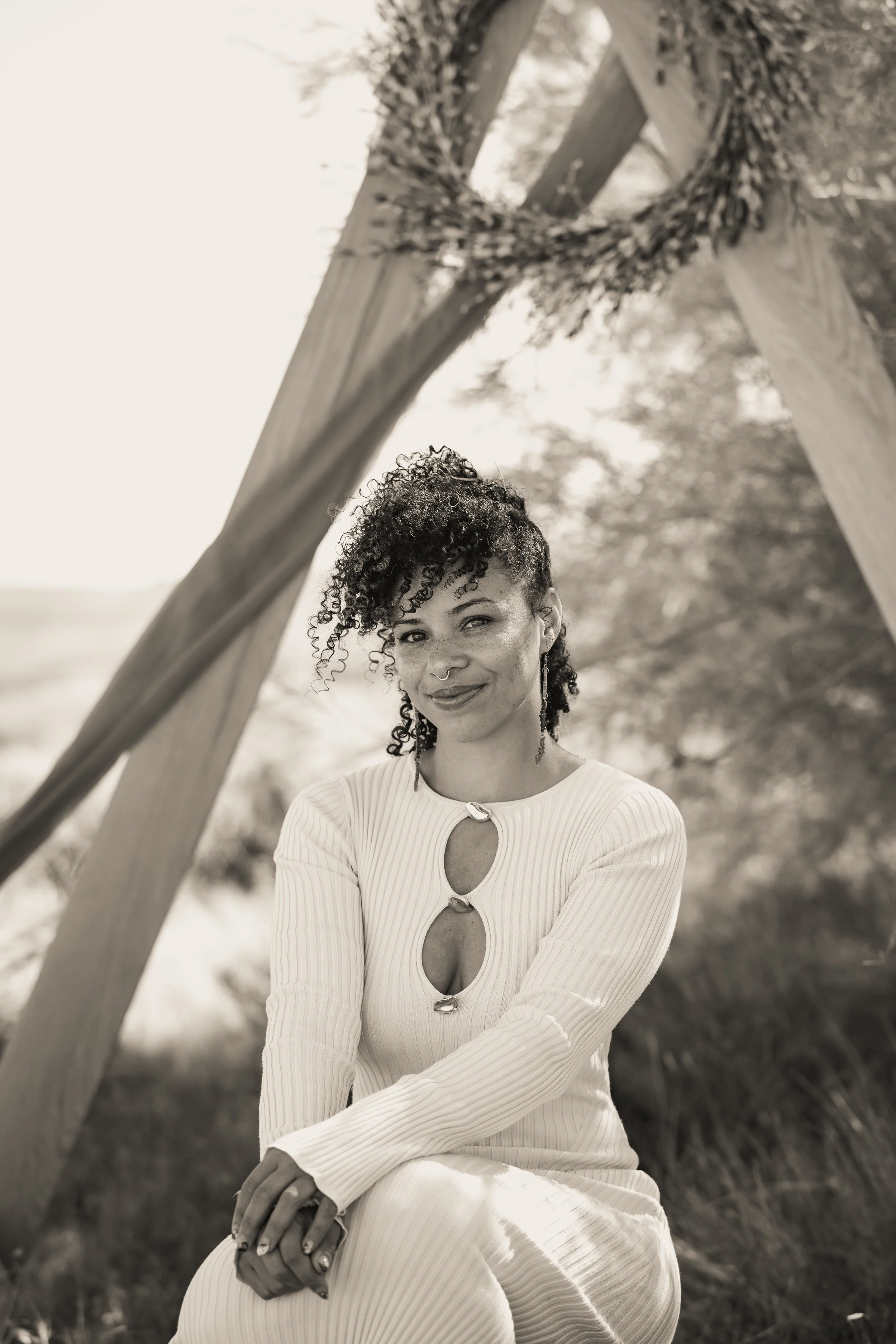 A woman with curly hair sitting outdoors in front of a wooden structure decorated with a wreath. She is wearing a long-sleeved, ribbed dress with a cut-out design and smiling slightly.