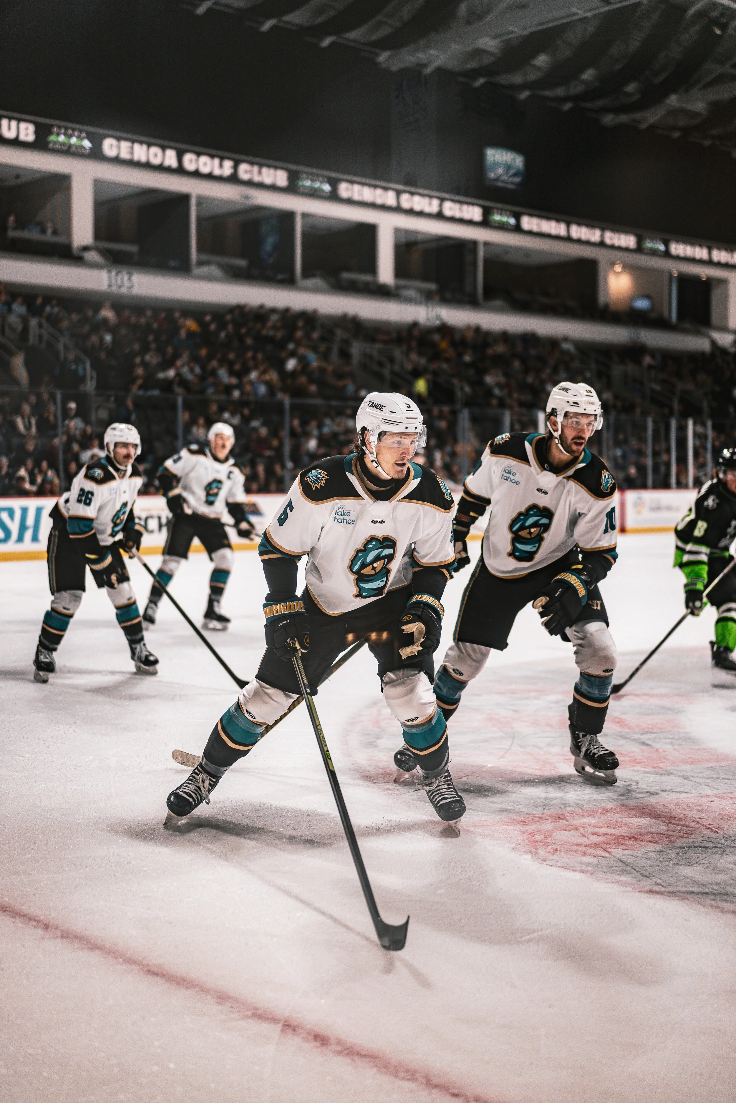 Hockey players in white and black uniforms skating on ice, with fans watching from the stands and advertisements on the boards.
