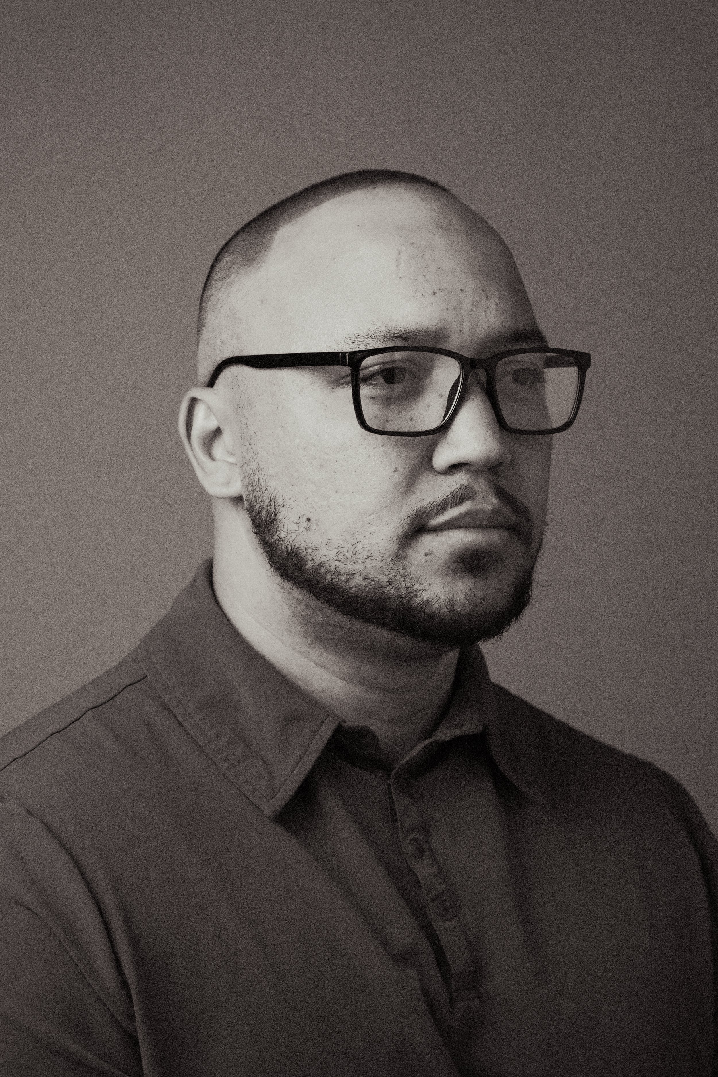 A black-and-white portrait of a man wearing glasses and a collared shirt, looking slightly to the side with a neutral expression.