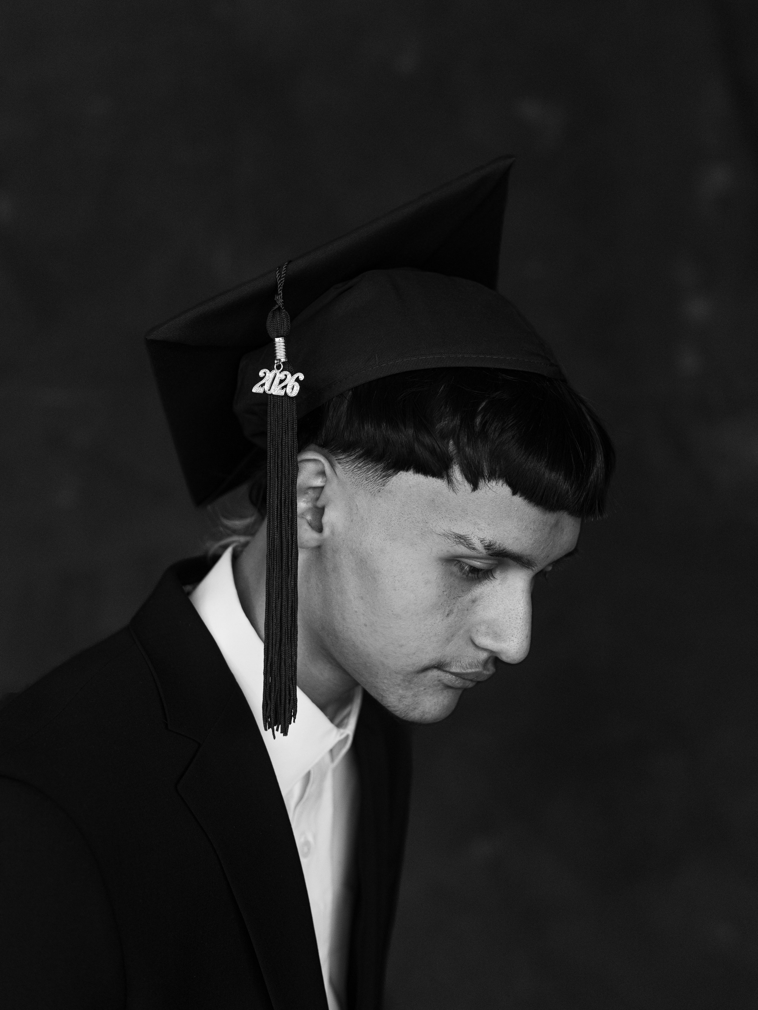 A young man in a graduation cap and gown looking down, against a dark background.