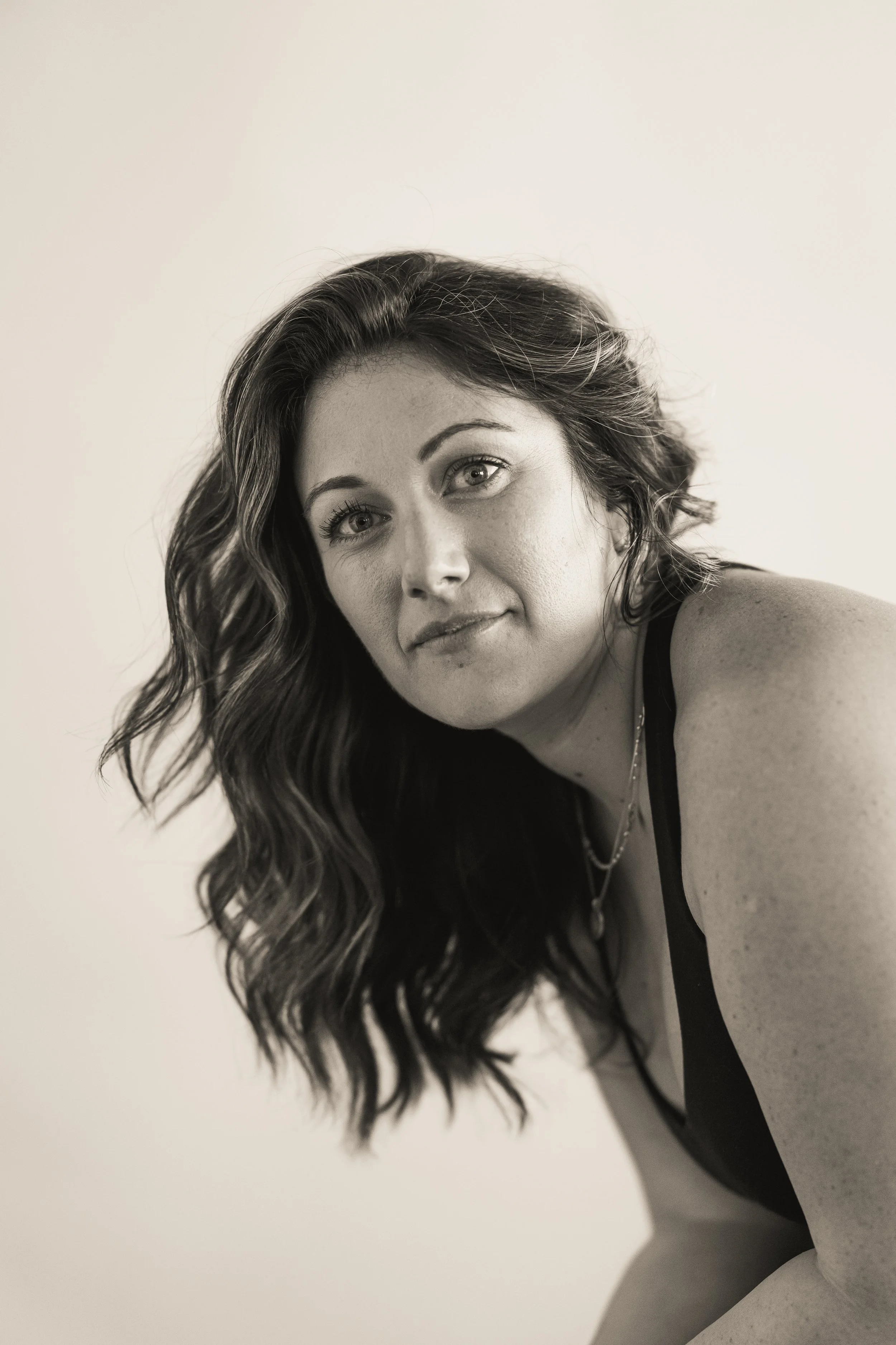 Black and white portrait of a woman with wavy hair wearing a sleeveless top, looking at the camera.