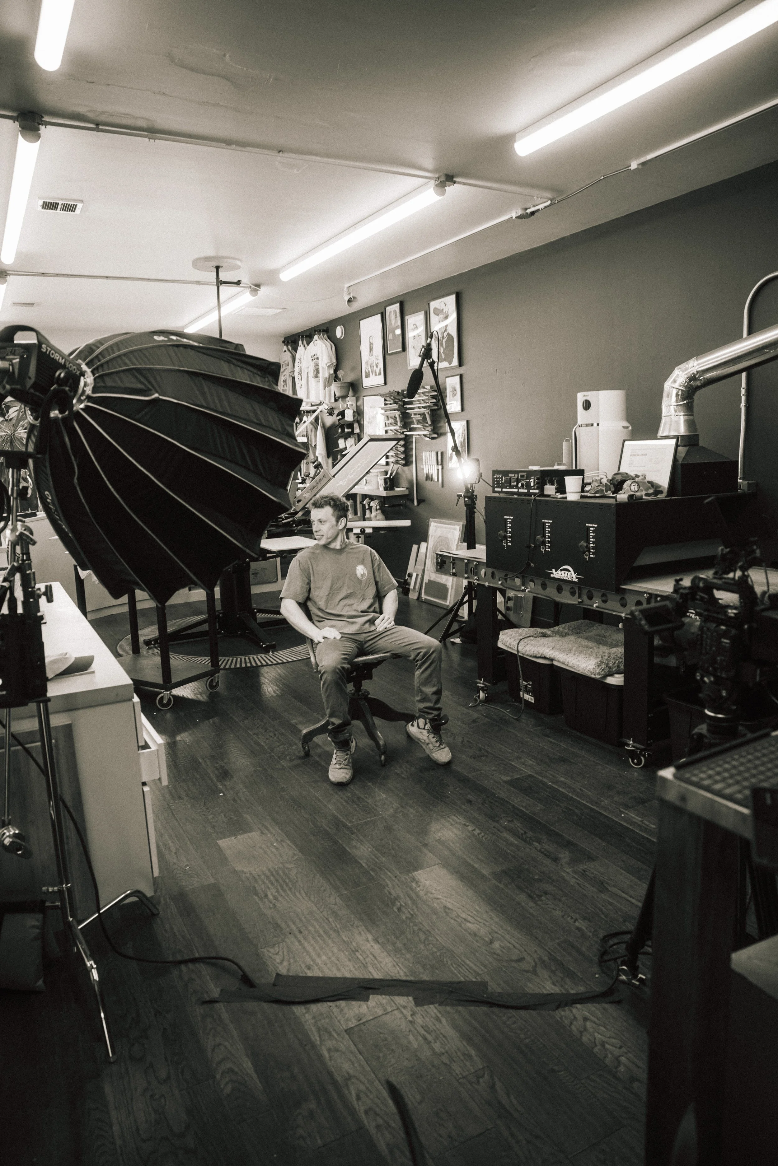 A man sits on a chair in a photography studio, surrounded by photography equipment including a large umbrella light, cameras, and lights. The studio has framed pictures and clothing on the wall.