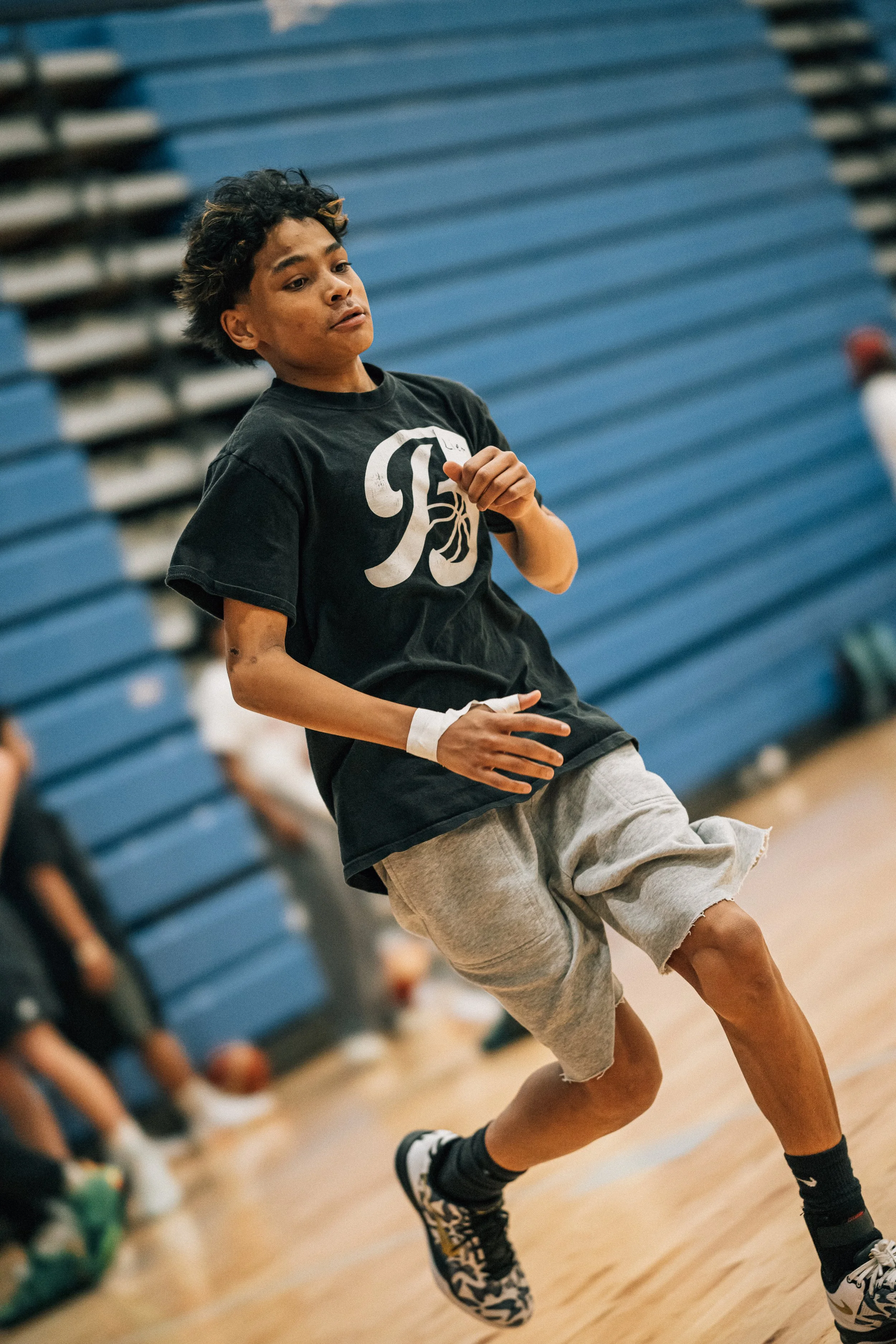 A young person with curly hair and a black t-shirt is jumping or running on a basketball court with blue bleachers in the background.