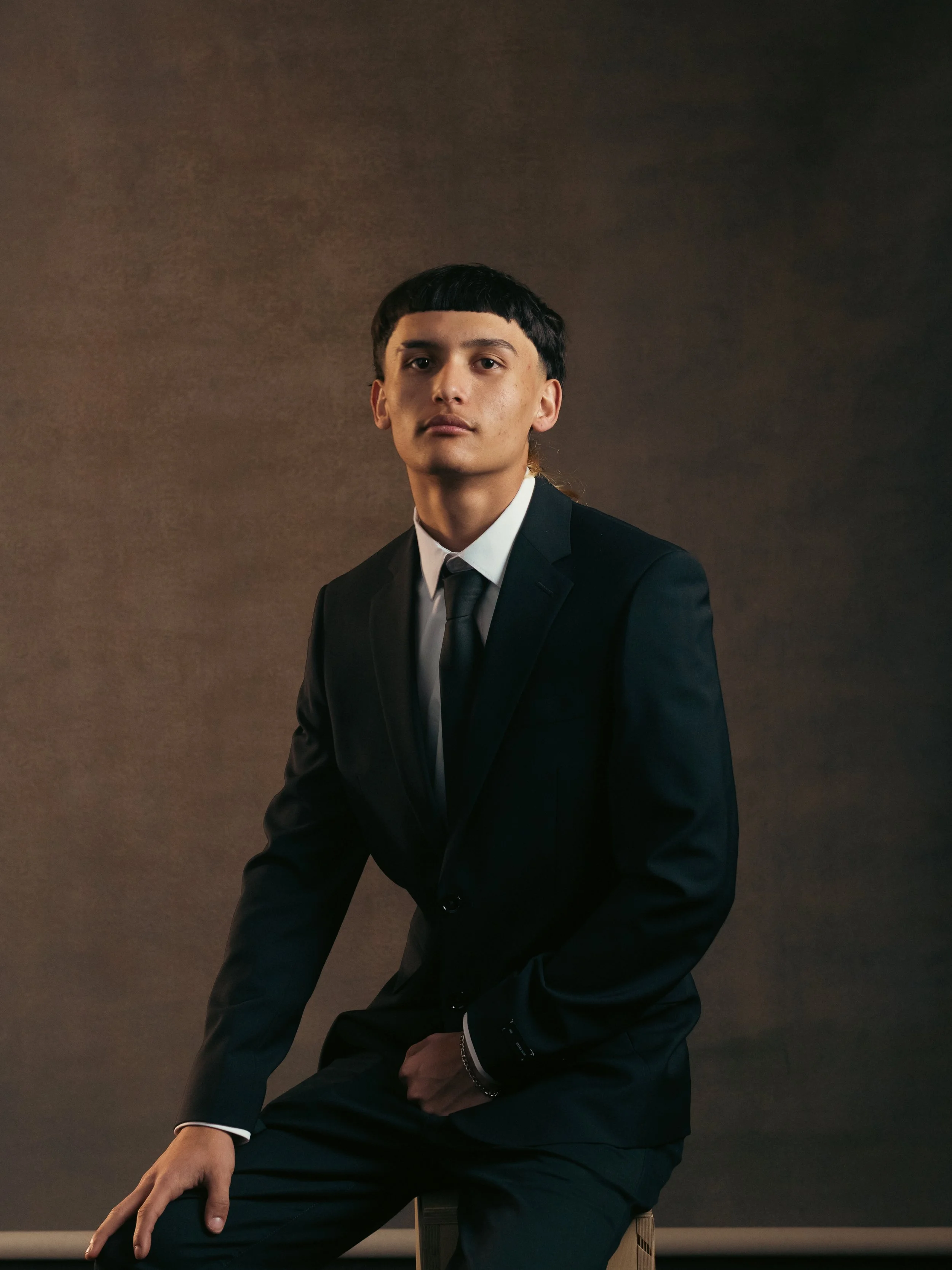 A young man with black hair and from the 80s wearing a black suit, white shirt, and black tie, sitting against a neutral backdrop.