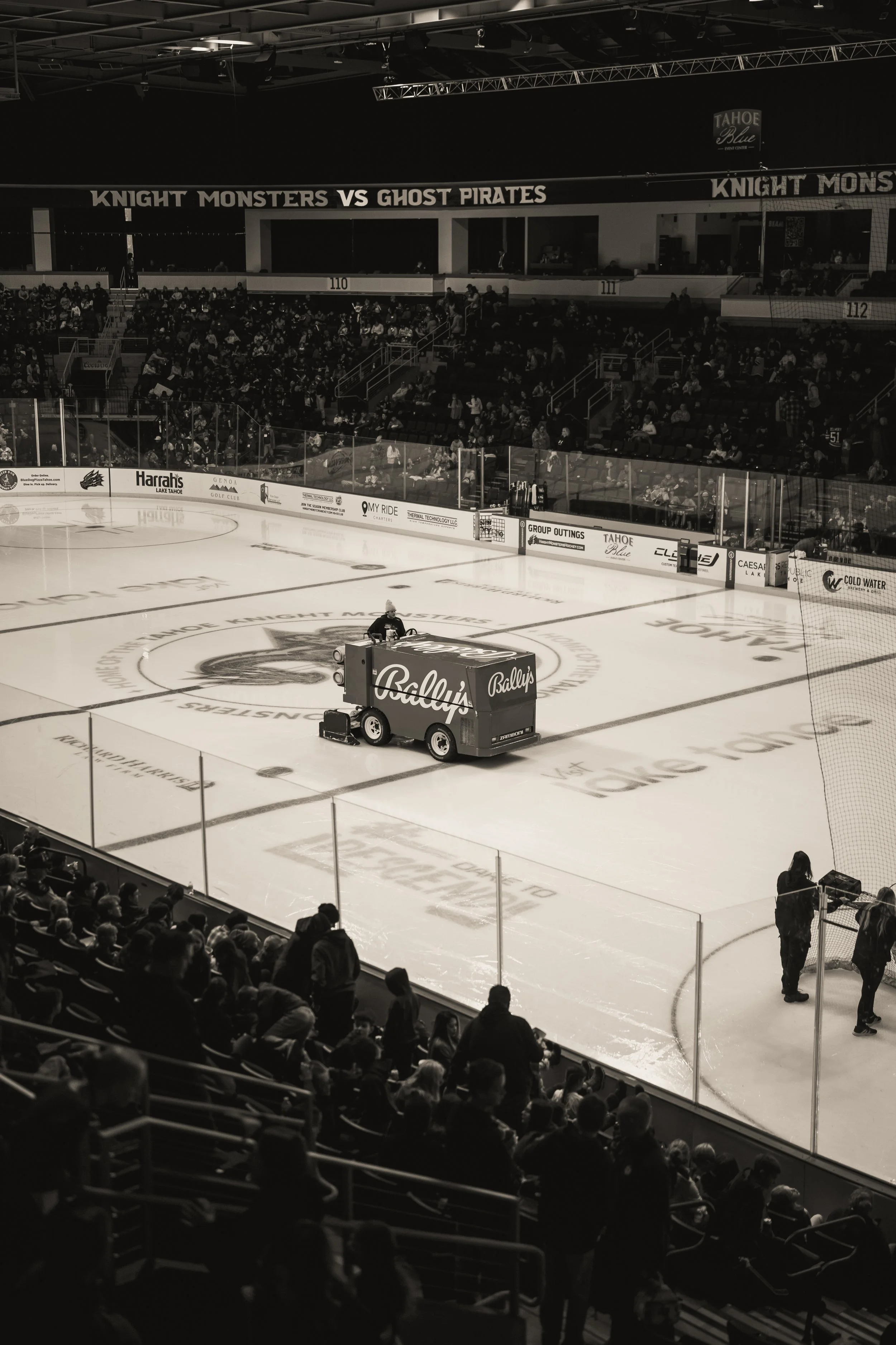 An indoor ice hockey arena with spectators seated and a Zamboni ice resurfacing machine on the ice. The arena features advertisements and personnel preparing the ice, with a banner reading 'Knight Monsters vs Ghost Pirates'.