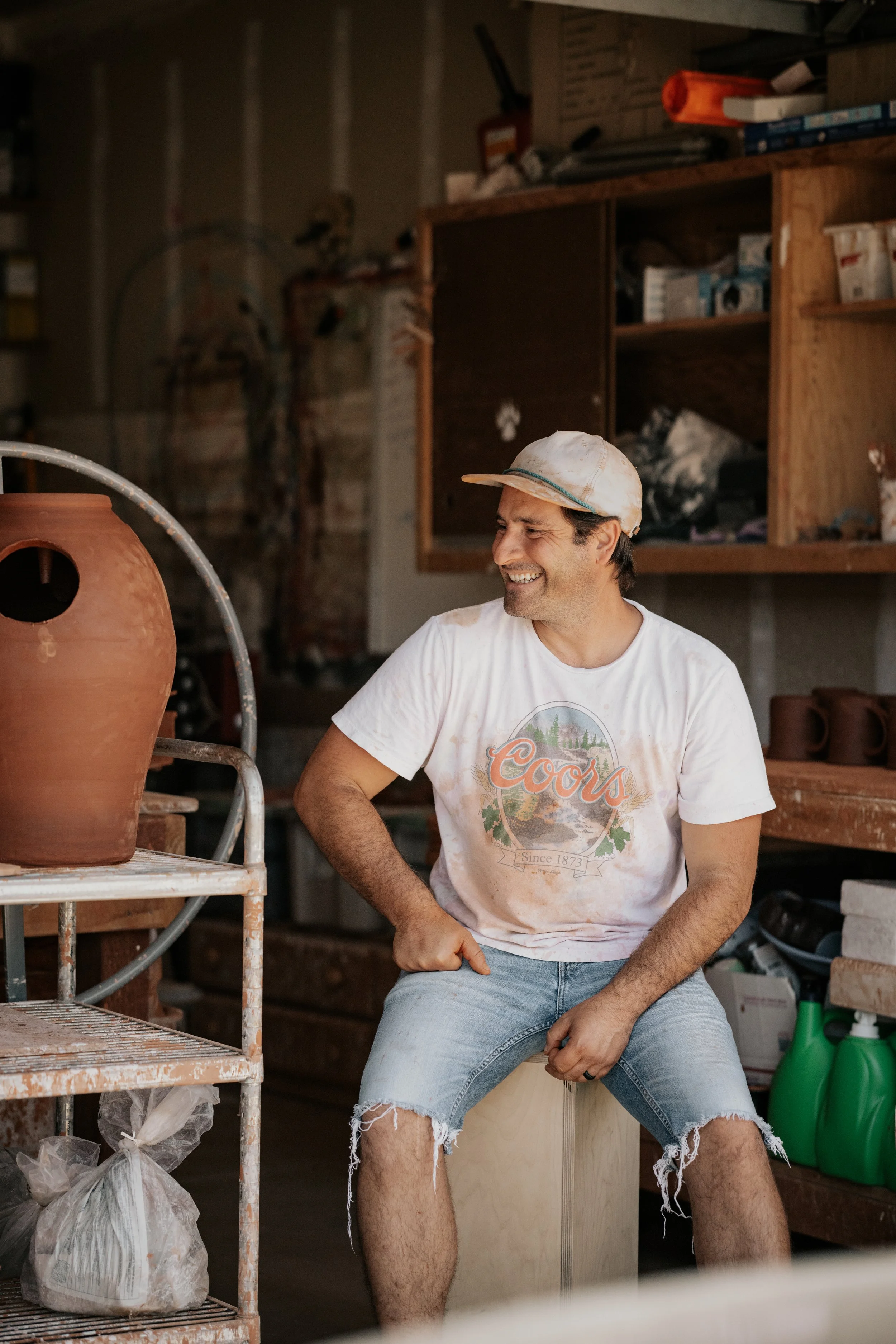 A man sitting on a wooden box smiling in a garage or workshop surrounded by shelves with tools and supplies, wearing a baseball cap, a dirty white T-shirt, and ripped denim shorts.