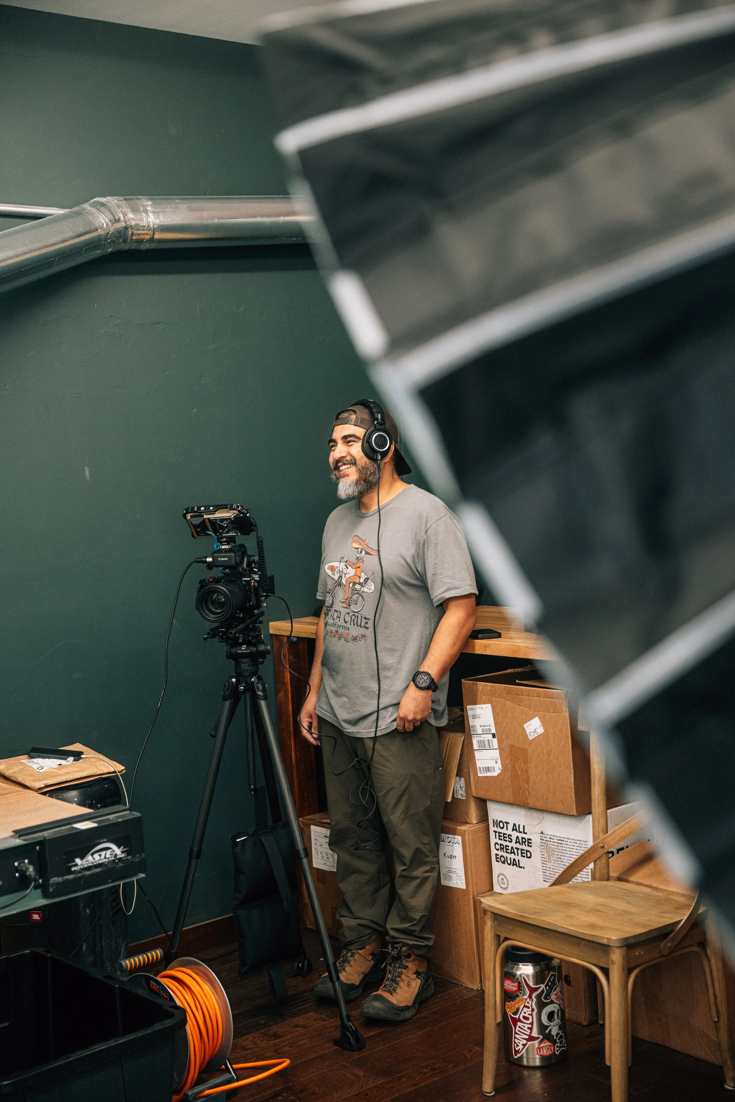 A man in gray t-shirt and khaki pants using a video camera on tripod, smiling, with headphones on, in a room with green wall and cardboard boxes.