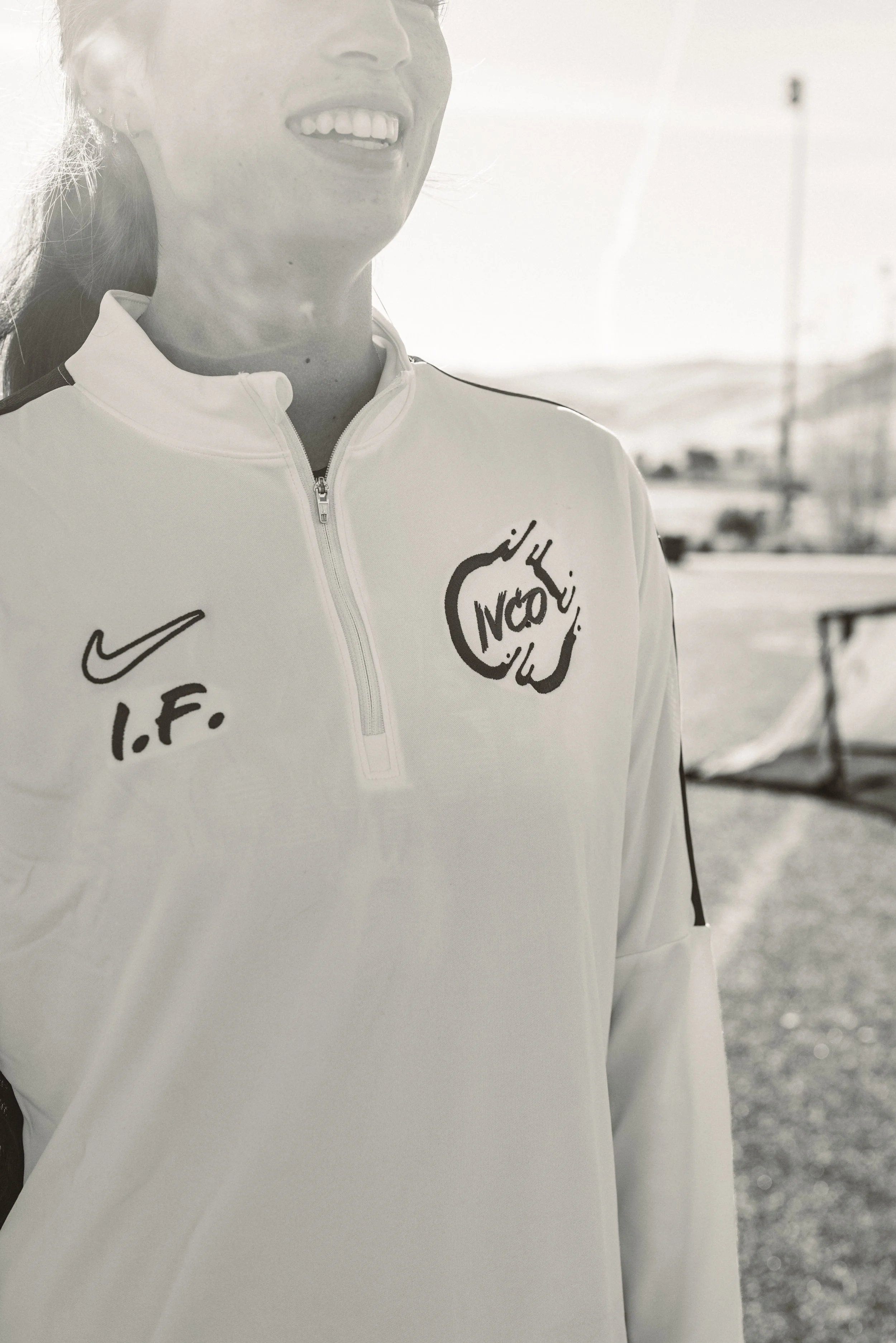 Close-up of a woman smiling in a white sports jacket with Nike logo and 'I.F.' initials, outdoors with a blurred background.