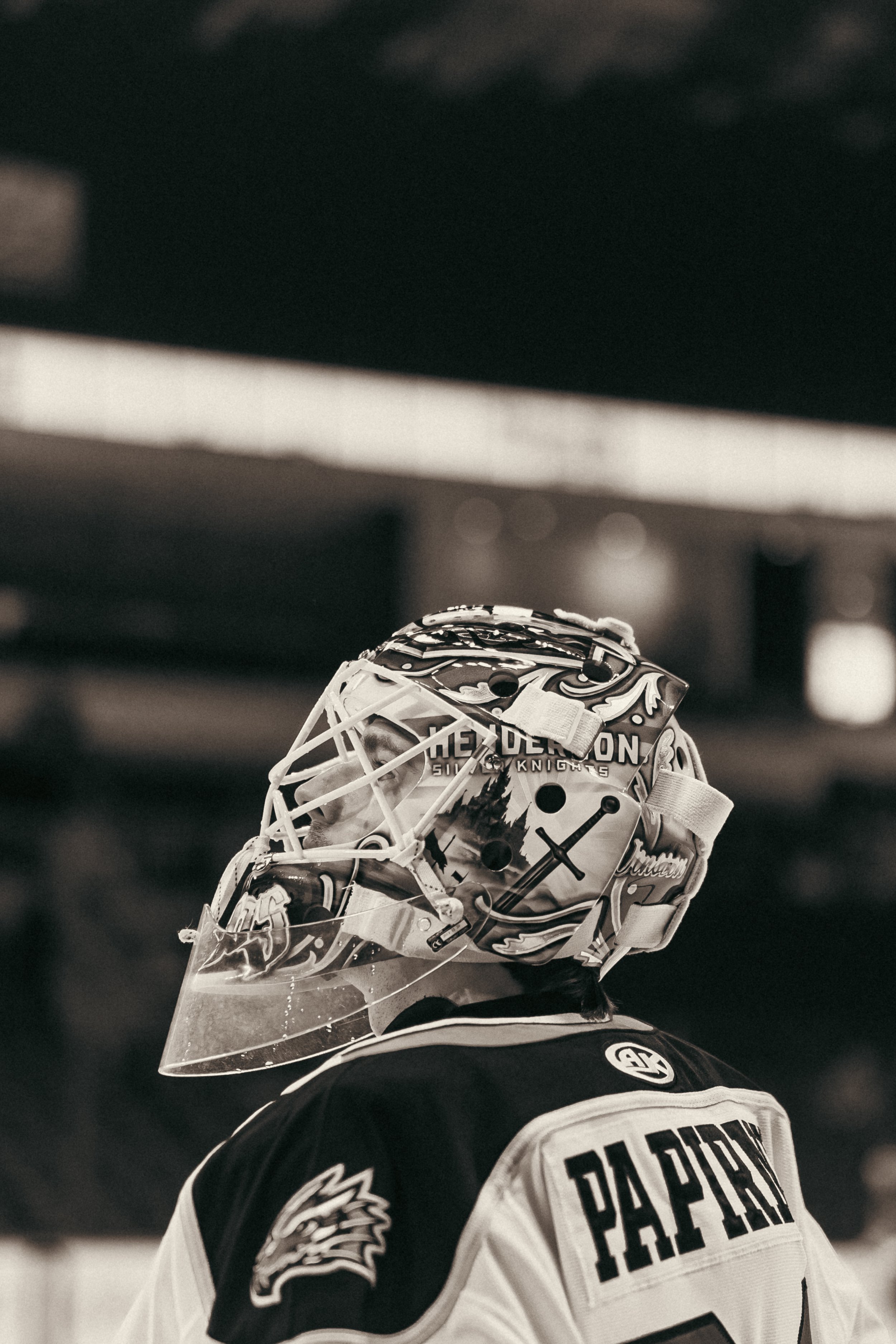 A hockey goalie wearing a detailed mask with various artworks and writing, and a jersey with the name Pappan on the back, standing on the ice in an indoor rink.