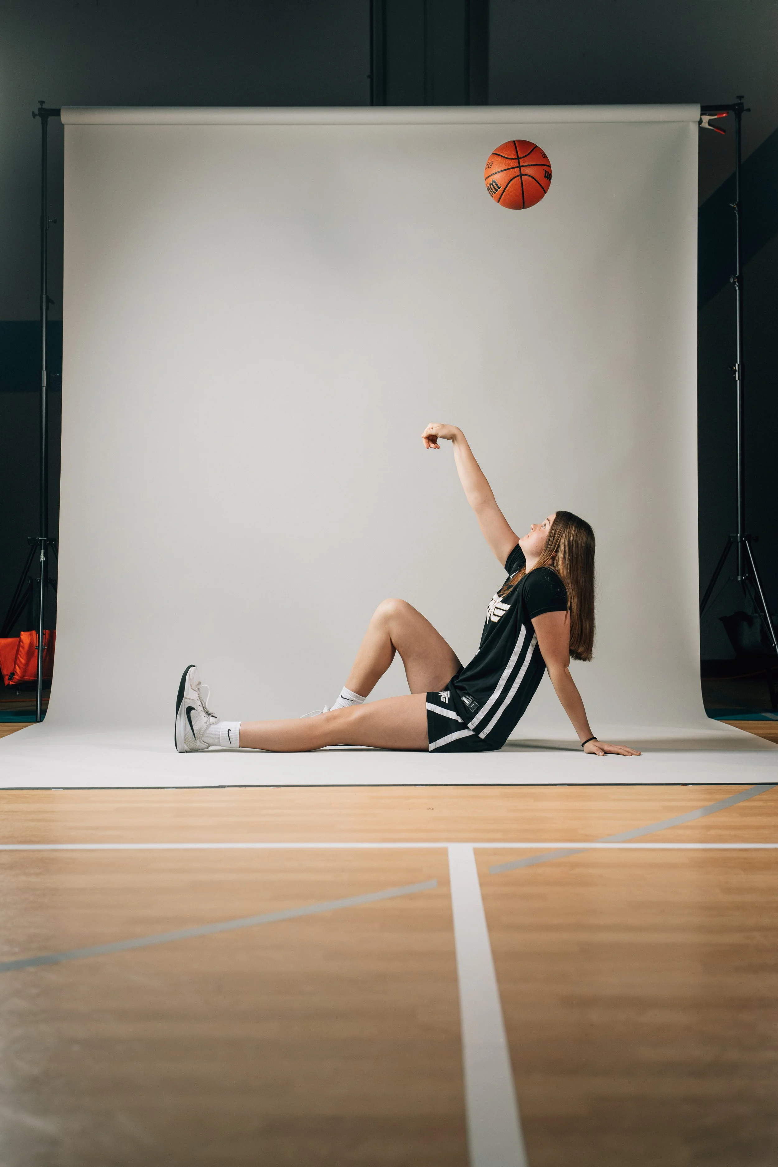 A young woman in black athletic wear and white sneakers sits on the floor of a gym, with a white backdrop behind her, as a basketball bounces in the air above her.
