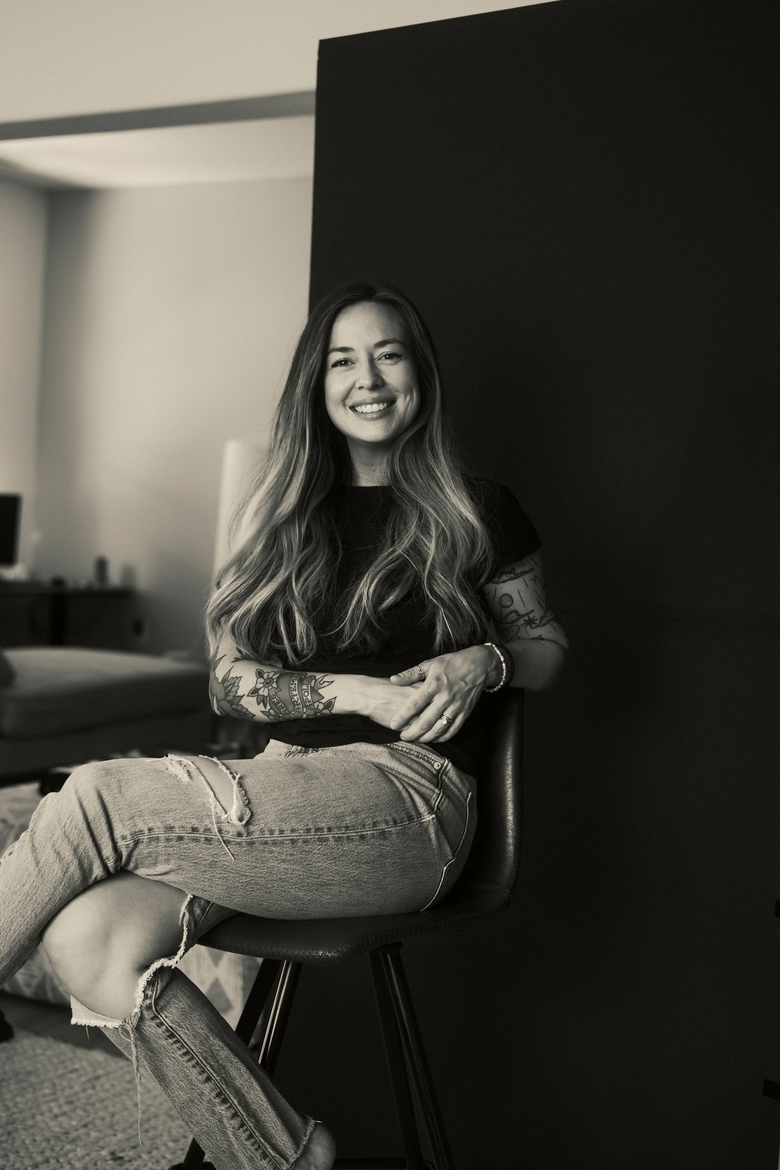 A woman with long, wavy hair and tattoos on her arms, smiling while sitting on a chair in a room with a bed and television in the background. The image is in black and white.