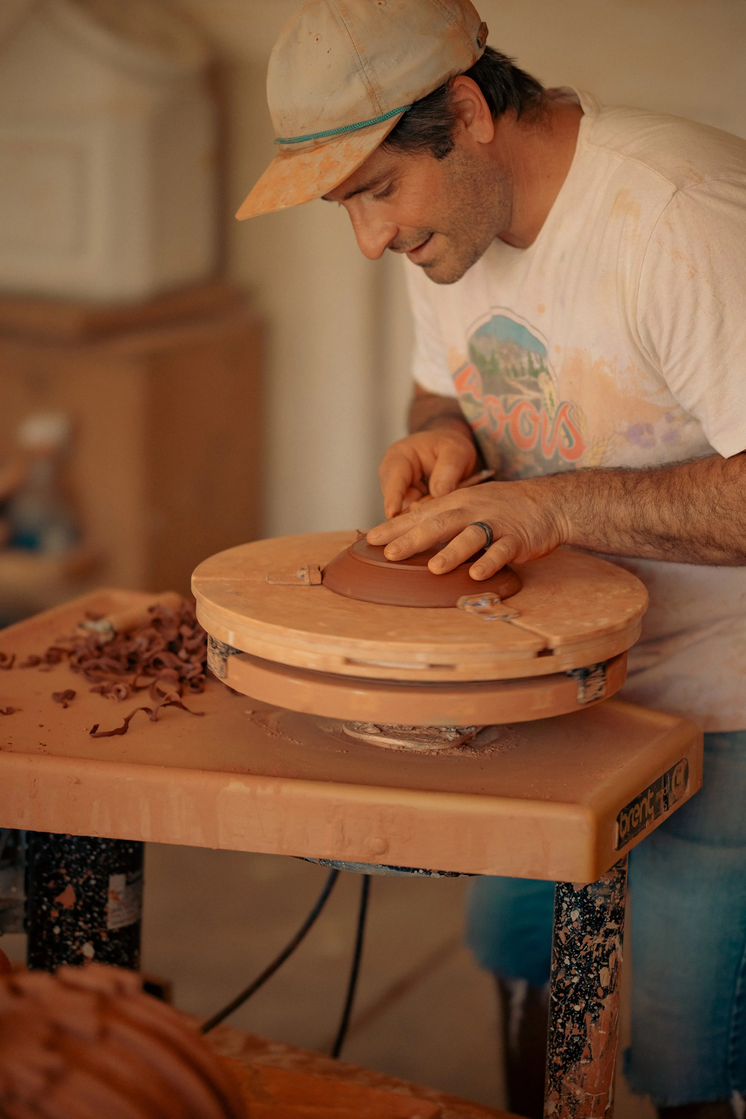 A man shaping clay on a pottery wheel, wearing a dirty baseball cap and a vintage t-shirt, in a pottery studio.
