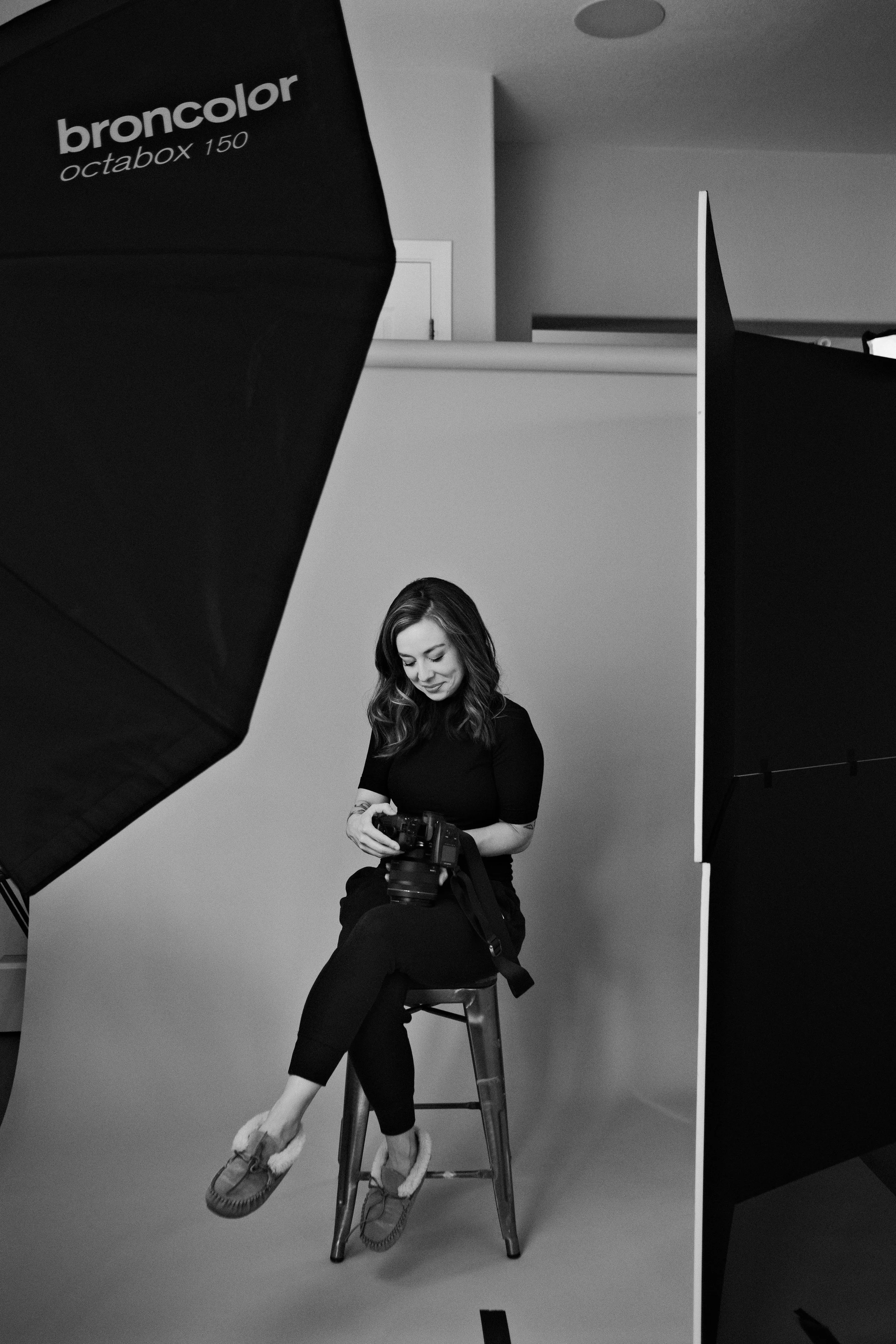 A woman sitting on a stool in a photography studio, holding a camera, surrounded by studio equipment, with a plain backdrop behind her.