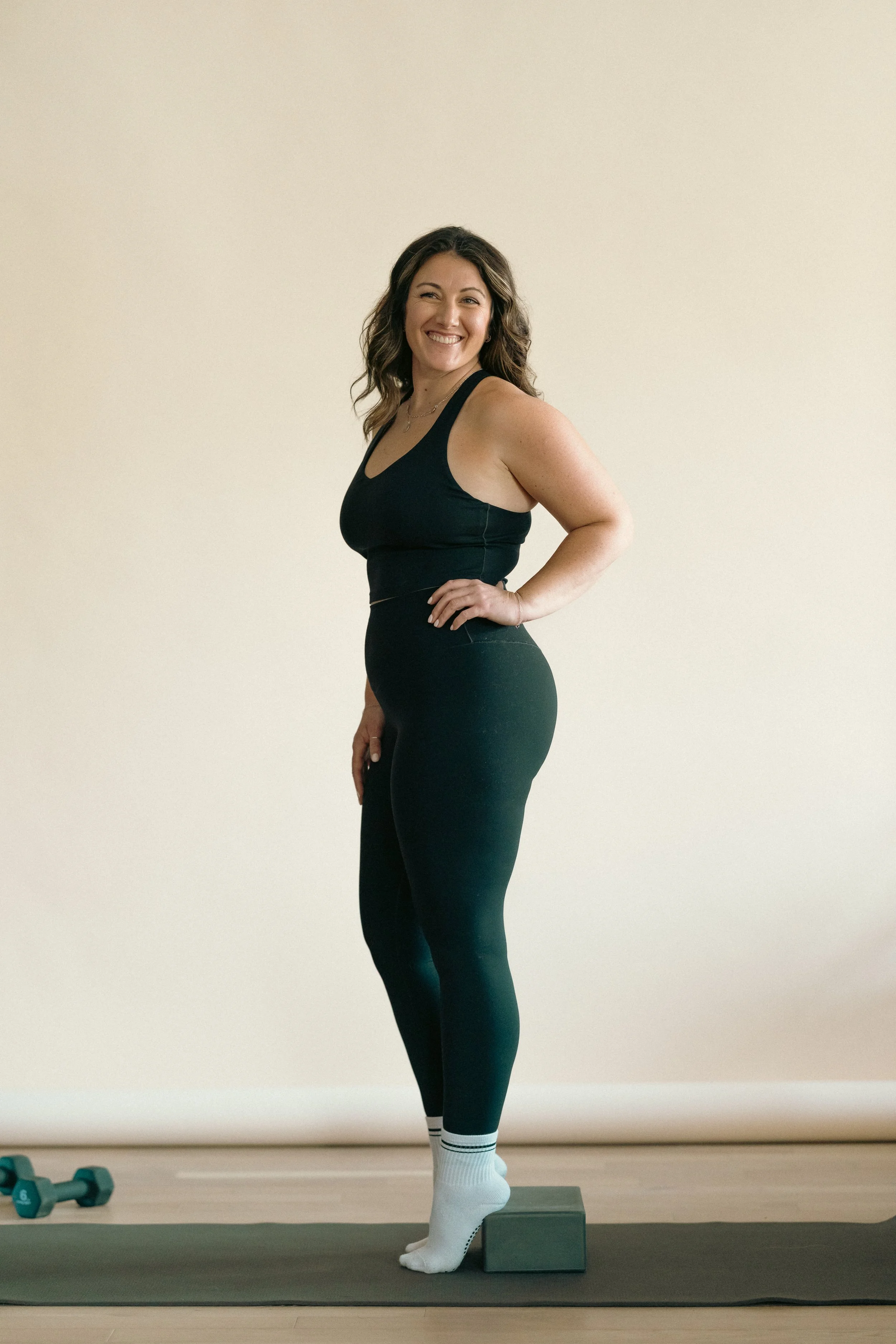 A woman in black workout clothes smiling and standing on a yoga block on a mat indoors.